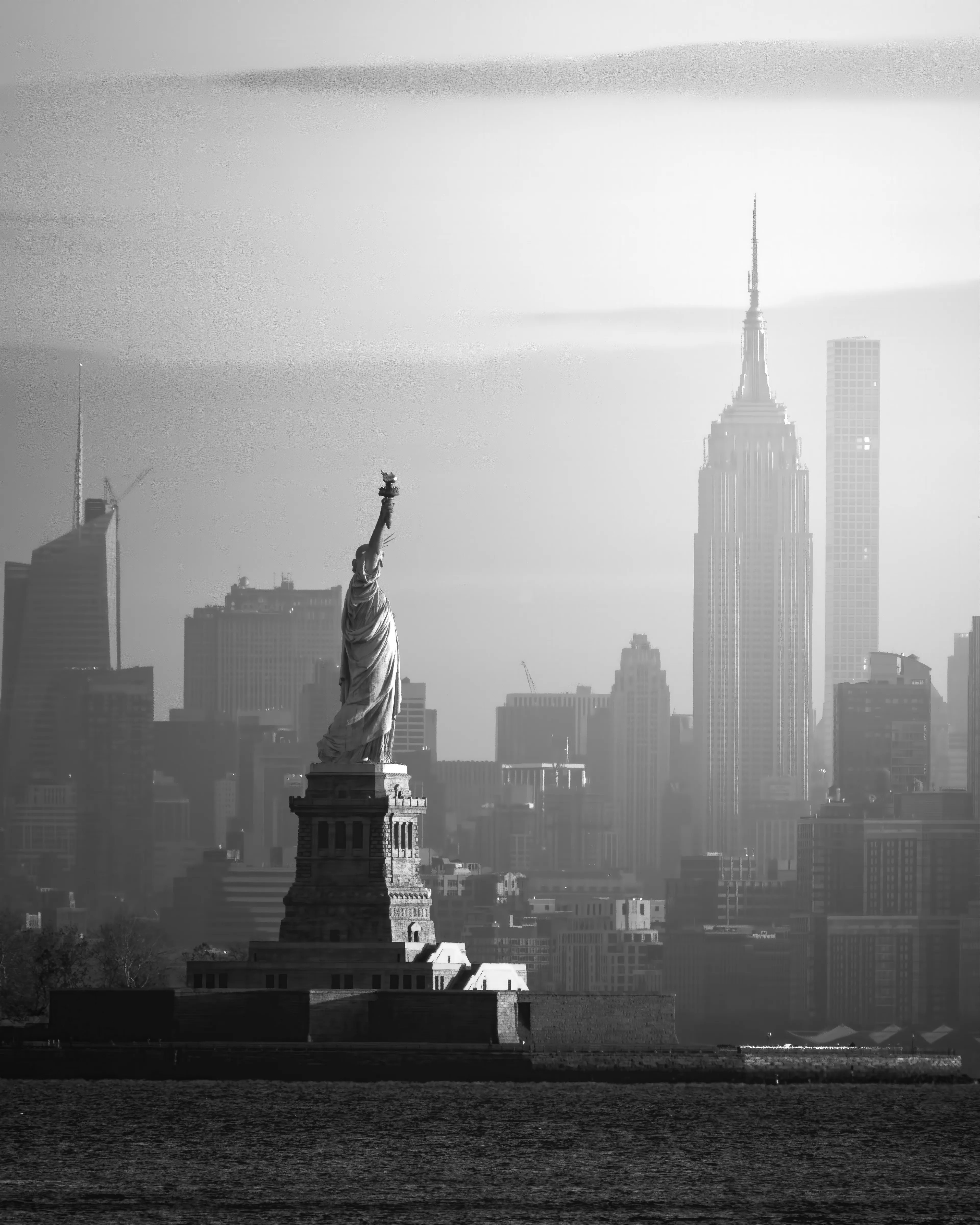 Early winter golden hour light illuminates the Statue Of Liberty.  In the morning glow, the atmospheric qualities  are accentuated by the distance from the vantage point to the Empire State Building and other skyline features are over 7 miles away.  Captured in color, but edited as a black and white image to create a timeless feel to the photo.  Light and shadow interplay create depth and highlight the directional movement of the morning light.