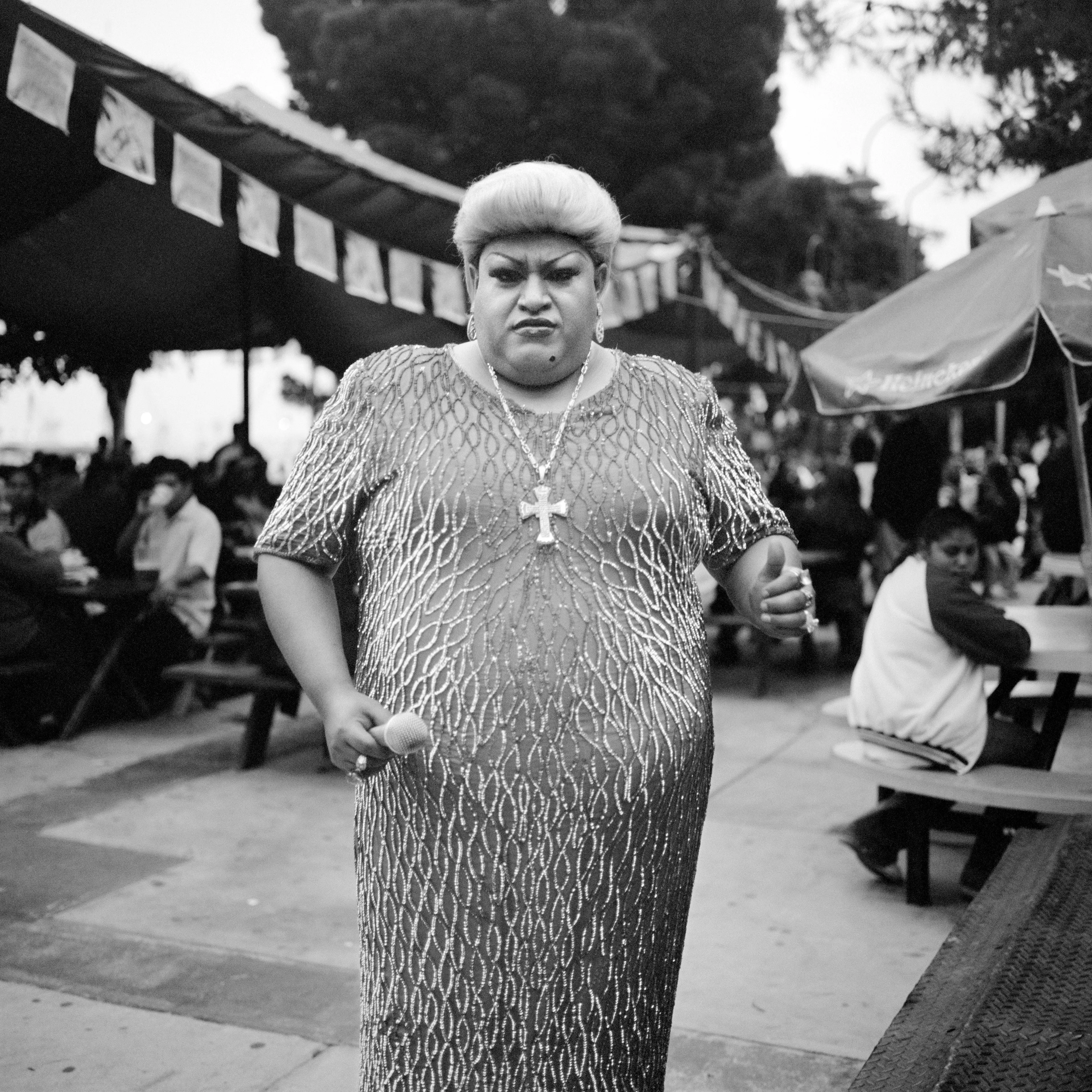 THE VILLAGE #20 PERFORMER IN A GOWN WEARING A CROSS by Deanna Templeton

This project started in 2002. A year after my mother passed away, a few years after my father passed. What I remember was one day I was thinking about my parents and feeling a little bit lonely, a little empty and decided to go for a drive. So I decided to drive out to my grandmother's house in San Pedro, which is about a 45 minute drive from where I live. I just wanted to reminisce, to re-explore the surroundings where I would go when I was a child. The liquor store I would walk to to get a Lipton Ice Tea and a bag of Frito’s, to the bakery where I swore all the cookies didn’t have sugar in them and then I drove over to Ports O’Call Village. As soon as I started to walk around I came across a stage with some Mariachis playing and it just hit me, I felt like I was home, I was with family. Over the next 5 years, whenever I could, I started going out there on the weekends. Getting reacquainted with the village and the locals. To shoot everything I wanted to remember.

Edition: 1 of 1

Commissioned by Obscura