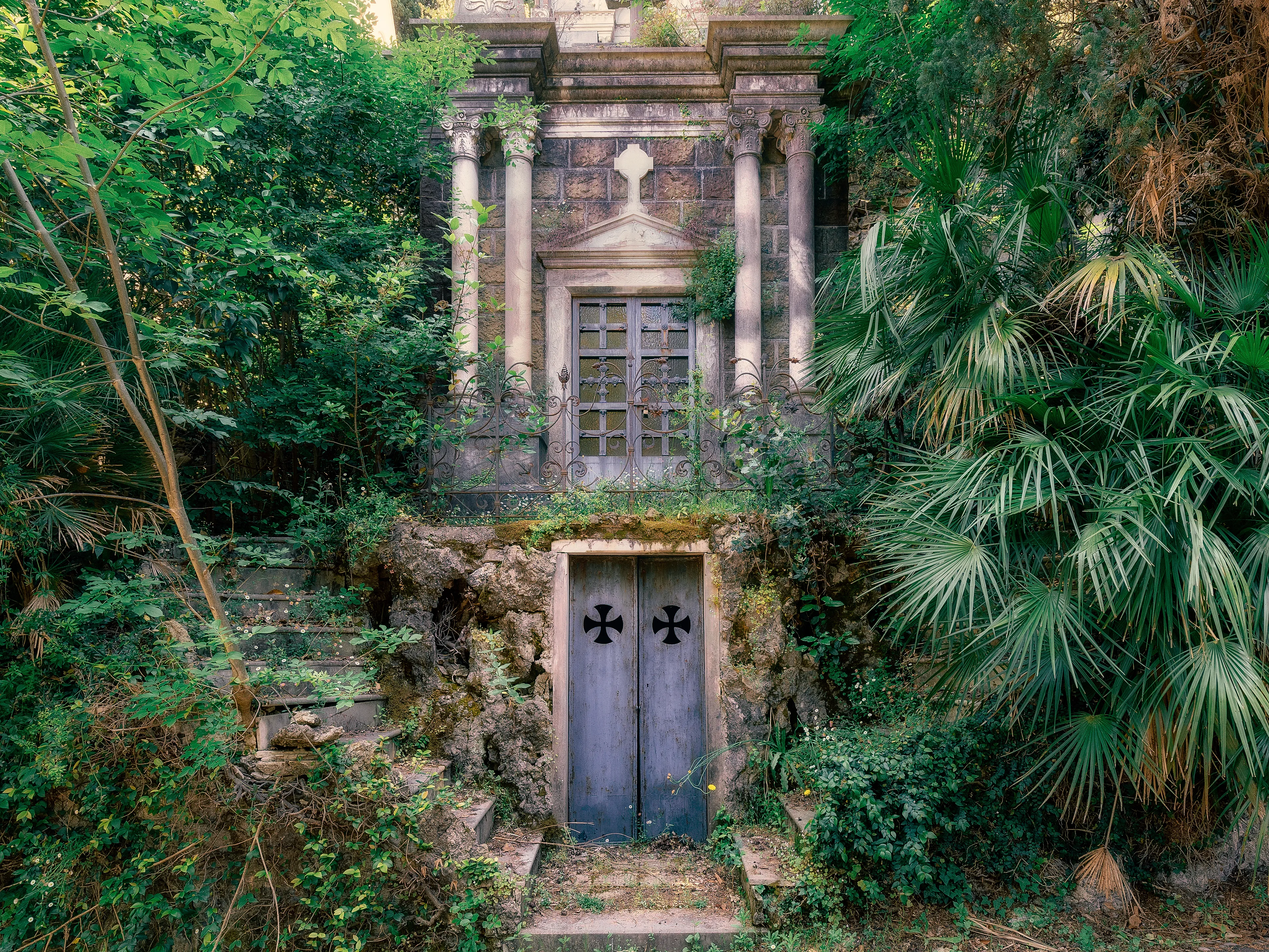 The Cimitero monumentale di Staglieno in Genoa, is one of the most beautiful and largest cemeteries in Europe.

It is known for incredibly detailed sculptures and monumental tombs.

Because of the age of some tombs and especially because of the size of the cemetery, there are also many forgotten places to discover. 

This old family tomb is located in a very remote part. The last persons were buried here in the 70's and it is an architectural beauty that is completed with a staircase that surrounds the tomb from both sides. 
Slowly but surely it is engulfed by tropical plants and one could think one has discovered a historical burial place in the jungle.