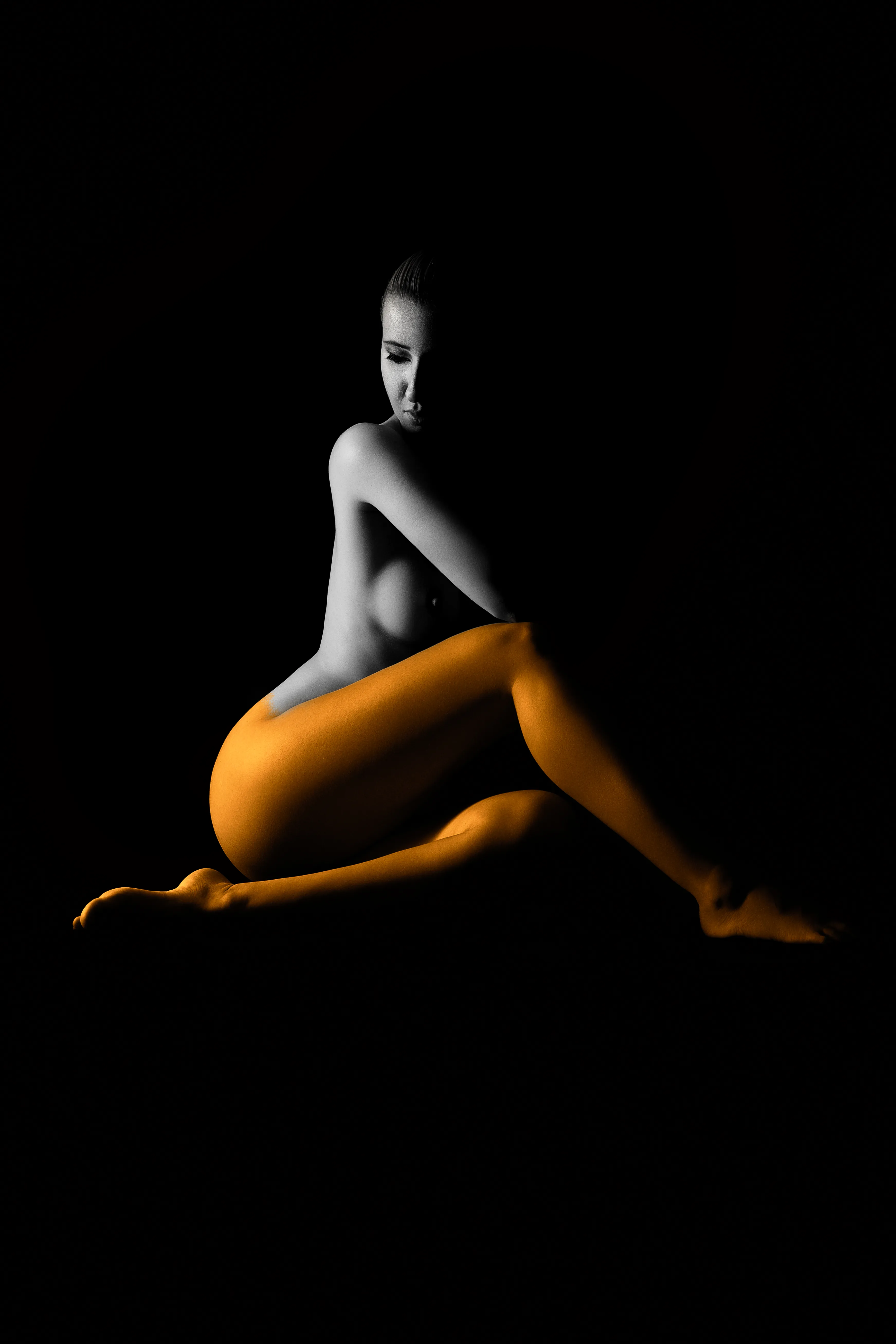 She is sitting into a shadow. She is half painted with orange.
Nude Art Photography.