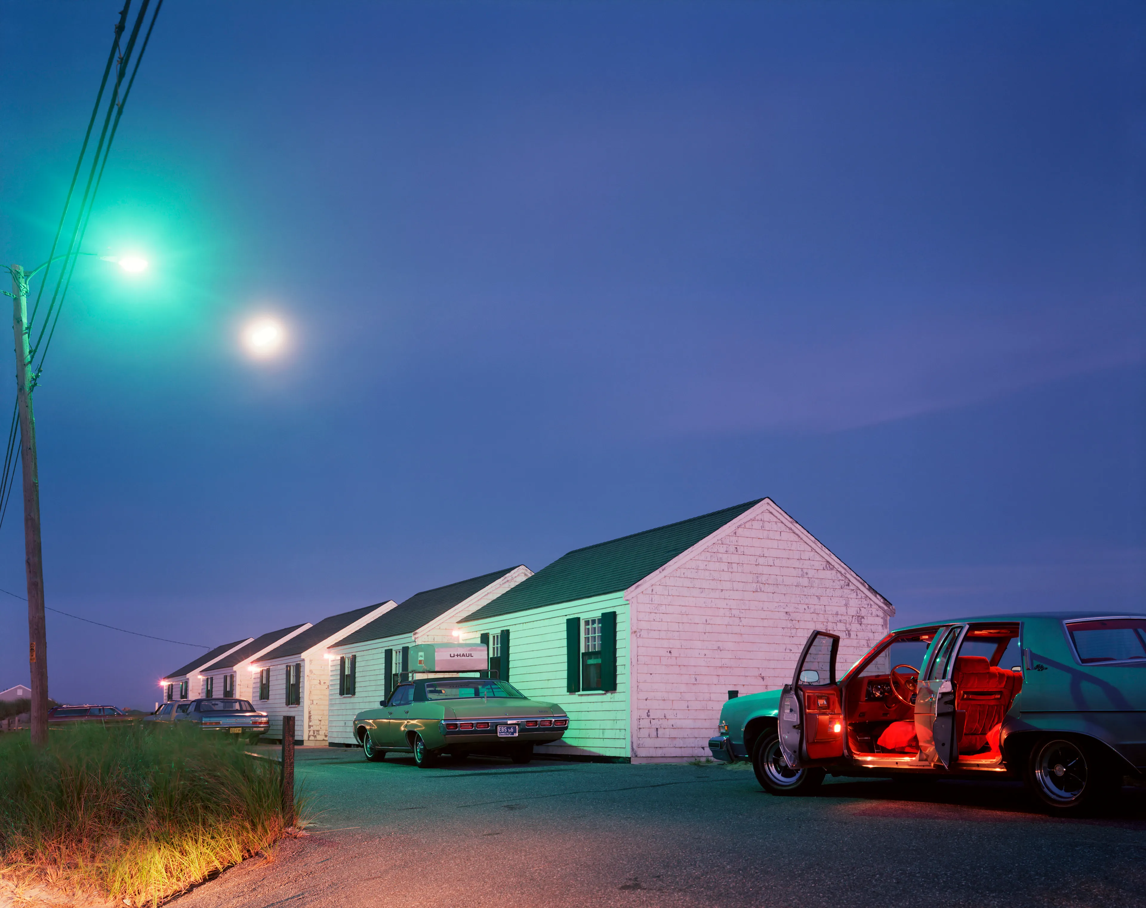 July 28, 1977: Cottages, Moon, Red Car Red Interior, Cars, Cottages, grass, street light, dusk, evening, “the blue hour” wires, uhaul, houses, night, telephone pole, car door, dusk.

"I took myself off to Provincetown on Cape Cod so I could summer with my family, and learn how to see in 8x10, in which the image is upside down and reversed, while beginning to learn to use Time in seconds, rather than in 1000th of a second. Well, Cape Cod is not 5th Avenue, but the light out there on that 60-mile-long sandbar in the Atlantic is unlike light on the mainland. Every molecule of moisture refracts light like a prism; while on the mainland there is a particle of dirt in every drop of atmosphere. Once I recognized that, then everything changed. Light, in all its intensity and tone, became my subject. Just as in 35mm, you can only photograph where you are and what you see there." - Joel Meyerowitz.

Edition: 1 of 1