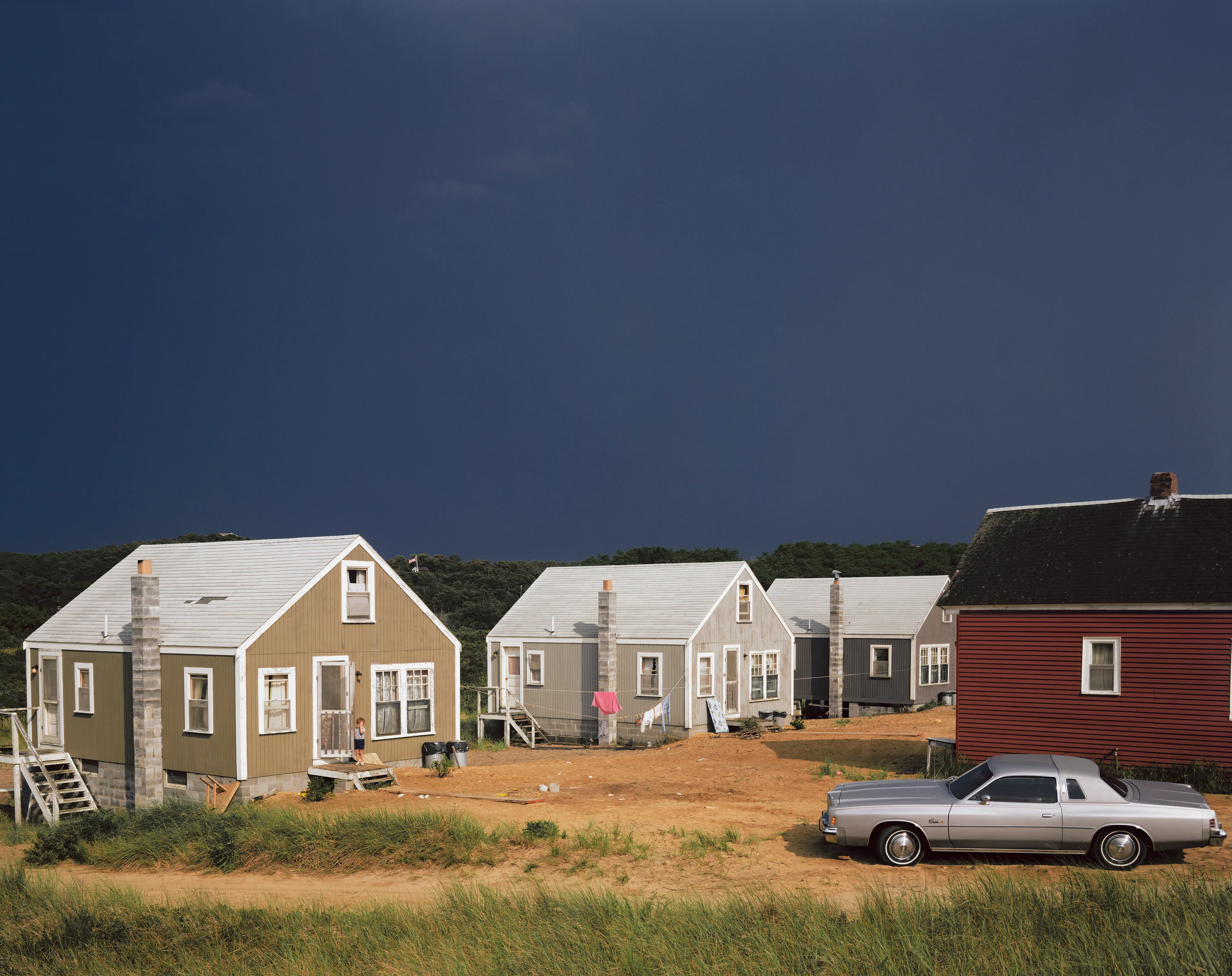 July 12: Pink Tower Dark Sky Cottages, Car, Kid, standing, storm, clothes line, laundry, houses, chimney, deck, porch, child.

"I took myself off to Provincetown on Cape Cod so I could summer with my family, and learn how to see in 8x10, in which the image is upside down and reversed, while beginning to learn to use Time in seconds, rather than in 1000th of a second. Well, Cape Cod is not 5th Avenue, but the light out there on that 60-mile-long sandbar in the Atlantic is unlike light on the mainland. Every molecule of moisture refracts light like a prism; while on the mainland there is a particle of dirt in every drop of atmosphere. Once I recognized that, then everything changed. Light, in all its intensity and tone, became my subject. Just as in 35mm, you can only photograph where you are and what you see there." - Joel Meyerowitz.

Edition: 1 of 1