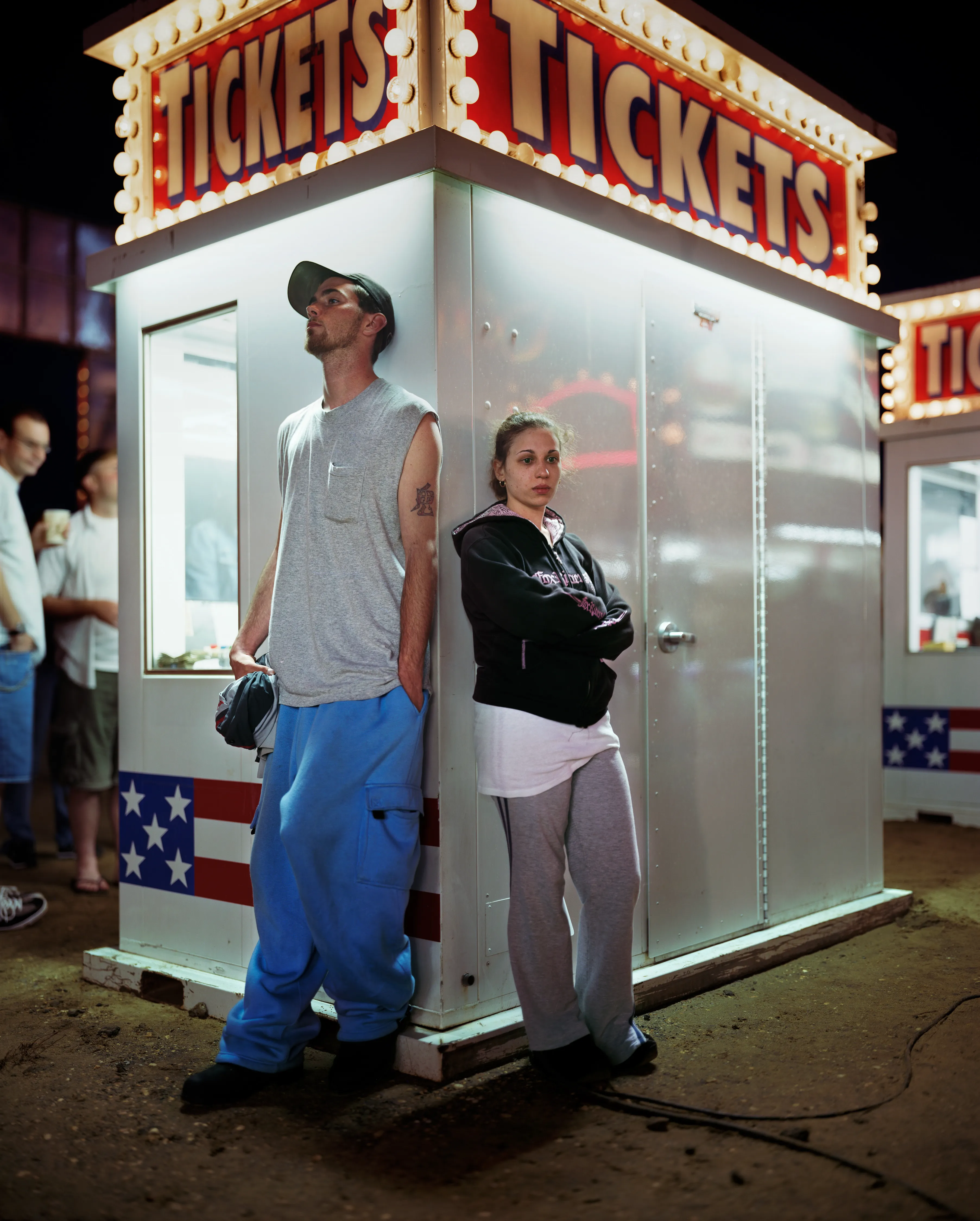 County fairs conjure ideas of tilt-a-whirls, stuffed animals, cotton candy, and blue ribbons. Rooted in local agricultural practices, fairs have been a ubiquitous part of American life for two centuries. In a reassuring way, they seem impervious to economic, technological, or fashion trends. For better or for worse, fairs look the same through the decades.

Though they are places to meet each other, there sure is a lot of loneliness at the fair. The series County Fair captures that duality: an adolescent boy looks up at the woman working the Hot Dog on a Stick stand as if she were a museum diorama; a little red haired girl haltingly befriends a robot; young couples, leaning and draped over each other, hide from the world in plain sight.


In my pictures, the dizzying atmosphere, colorfully decorated to provide amusement and distraction, is imbued with sadness and longing. These feelings, which permeate society, are laid bare at the fair. Sadness and longing circulate, intersect and incubate in the confined space between the ticket booths and the craft barns. People come out to participate and show, to see and be seen. Ultimately, connection remains elusive. 

