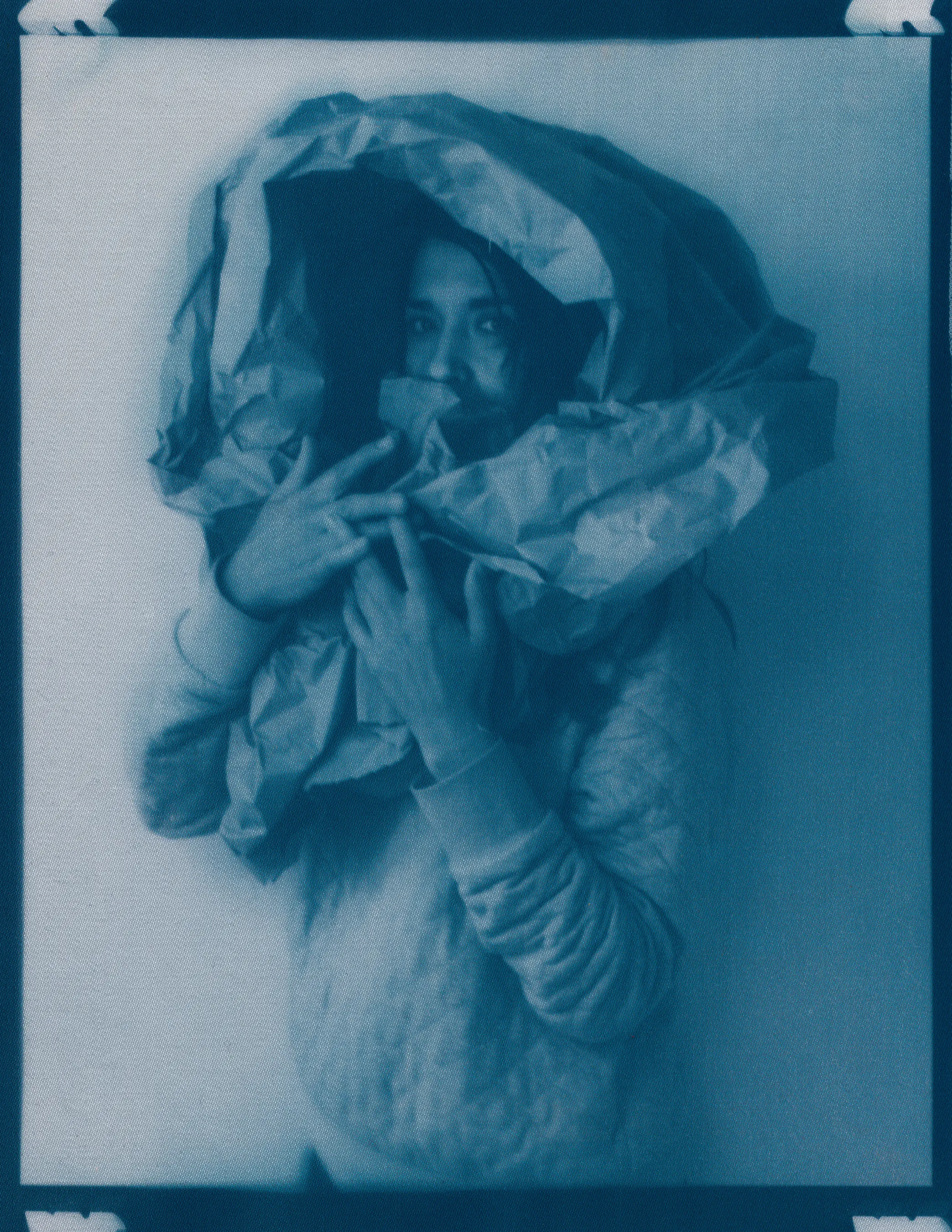 In the spring of 2020, panic surrounding COVID-19 erupted and mandatory shelter in place orders went into effect, forcing me to abandon long-planned portrait shoots, travel, and work in progress in my art studio.  I found myself in a 340 sq ft apartment in a Bay-area neighborhood that was emptying by the day.  The buzzing silence was profound. Two weeks into isolation, my drive to connect with others and make portraits only amplified.  With no safe way to do so, I spontaneously turned the camera on myself to create a portrait.  I printed it as a cyanotype, a simple nineteenth century photographic process that was only feasible due to the basic materials I happened to have on hand—a budget printer, transparency film and a package of fraying sheets of cotton pretreated with cyanotype chemicals.  At first, I exposed the prints in the unpredictable spring sunlight coming through my third floor apartment window.  As the seasons changed, I went on to make them in a garden, on the top of a car, or a patch of wobbly concrete tiles.  I rinsed them in water and varying shades of blue emerged.  I created a daily self-portrait using this technique for more than a year. 

I made most of these self-portraits inside various studio apartments that I lived in alone.  To make the earliest pictures in the series, I had to move a couch every day to create space against a white wall near a window.  I took others in fleeting spaces while traveling—in a guest room, in a medical examination room, during a pause in the wilderness, and later against the wall of an old California bungalow sandwiched between the mountains and the sea.  In the beginning, I often tried to cover as much of my body and face as possible as a commentary on my fear of the virus and my efforts to guard against it.  My armor and props ranged from common household items—potholders, tinfoil, dish towels, bedsheets, and toilet paper— to more telling evidence of the unusual consumption that resulted from being stuck indoors indefinitely— Amazon packaging, takeout bags and trash leftover from groceries purchased while wearing rubber gloves and sterilized in whatever way  was possible and later consumed.  These items shifted as the duration of the pandemic blurred into an unknown stretch of time. The portraits became less about those initial fears and more about confronting the boredom, anxiety, grief, and fatigue of living in indefinite isolation during a global pandemic. 

In total, I made these portraits for 377 consecutive days.  I made the first portrait on March 30th, 2020, a day when my head was raging in pain, my throat was constricted, and the fear that I might have contracted COVID hung over me before testing was easily accessible.  I made the last portrait on April 10th, 2021, the day I received my 2nd vaccination.  The days, weeks, and months in between feel like a fever dream—frayed and flickering in multitudes of blue.

2021_0213