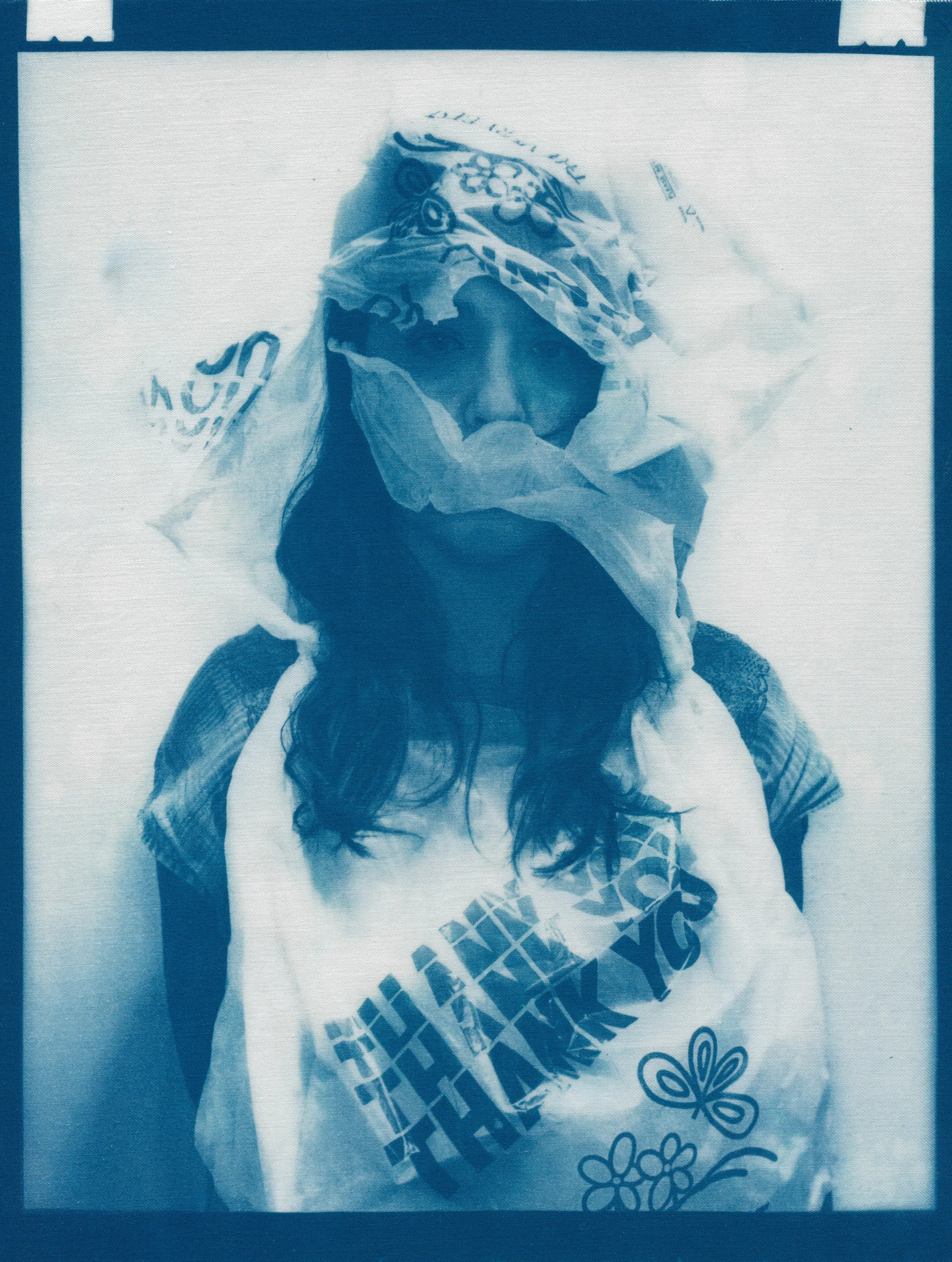 In the spring of 2020, panic surrounding COVID-19 erupted and mandatory shelter in place orders went into effect, forcing me to abandon long-planned portrait shoots, travel, and work in progress in my art studio.  I found myself in a 340 sq ft apartment in a Bay-area neighborhood that was emptying by the day.  The buzzing silence was profound. Two weeks into isolation, my drive to connect with others and make portraits only amplified.  With no safe way to do so, I spontaneously turned the camera on myself to create a portrait.  I printed it as a cyanotype, a simple nineteenth century photographic process that was only feasible due to the basic materials I happened to have on hand—a budget printer, transparency film and a package of fraying sheets of cotton pretreated with cyanotype chemicals.  At first, I exposed the prints in the unpredictable spring sunlight coming through my third floor apartment window.  As the seasons changed, I went on to make them in a garden, on the top of a car, or a patch of wobbly concrete tiles.  I rinsed them in water and varying shades of blue emerged.  I created a daily self-portrait using this technique for more than a year. 

I made most of these self-portraits inside various studio apartments that I lived in alone.  To make the earliest pictures in the series, I had to move a couch every day to create space against a white wall near a window.  I took others in fleeting spaces while traveling—in a guest room, in a medical examination room, during a pause in the wilderness, and later against the wall of an old California bungalow sandwiched between the mountains and the sea.  In the beginning, I often tried to cover as much of my body and face as possible as a commentary on my fear of the virus and my efforts to guard against it.  My armor and props ranged from common household items—potholders, tinfoil, dish towels, bedsheets, and toilet paper— to more telling evidence of the unusual consumption that resulted from being stuck indoors indefinitely— Amazon packaging, takeout bags and trash leftover from groceries purchased while wearing rubber gloves and sterilized in whatever way  was possible and later consumed.  These items shifted as the duration of the pandemic blurred into an unknown stretch of time. The portraits became less about those initial fears and more about confronting the boredom, anxiety, grief, and fatigue of living in indefinite isolation during a global pandemic. 

In total, I made these portraits for 377 consecutive days.  I made the first portrait on March 30th, 2020, a day when my head was raging in pain, my throat was constricted, and the fear that I might have contracted COVID hung over me before testing was easily accessible.  I made the last portrait on April 10th, 2021, the day I received my 2nd vaccination.  The days, weeks, and months in between feel like a fever dream—frayed and flickering in multitudes of blue.

2020_0914