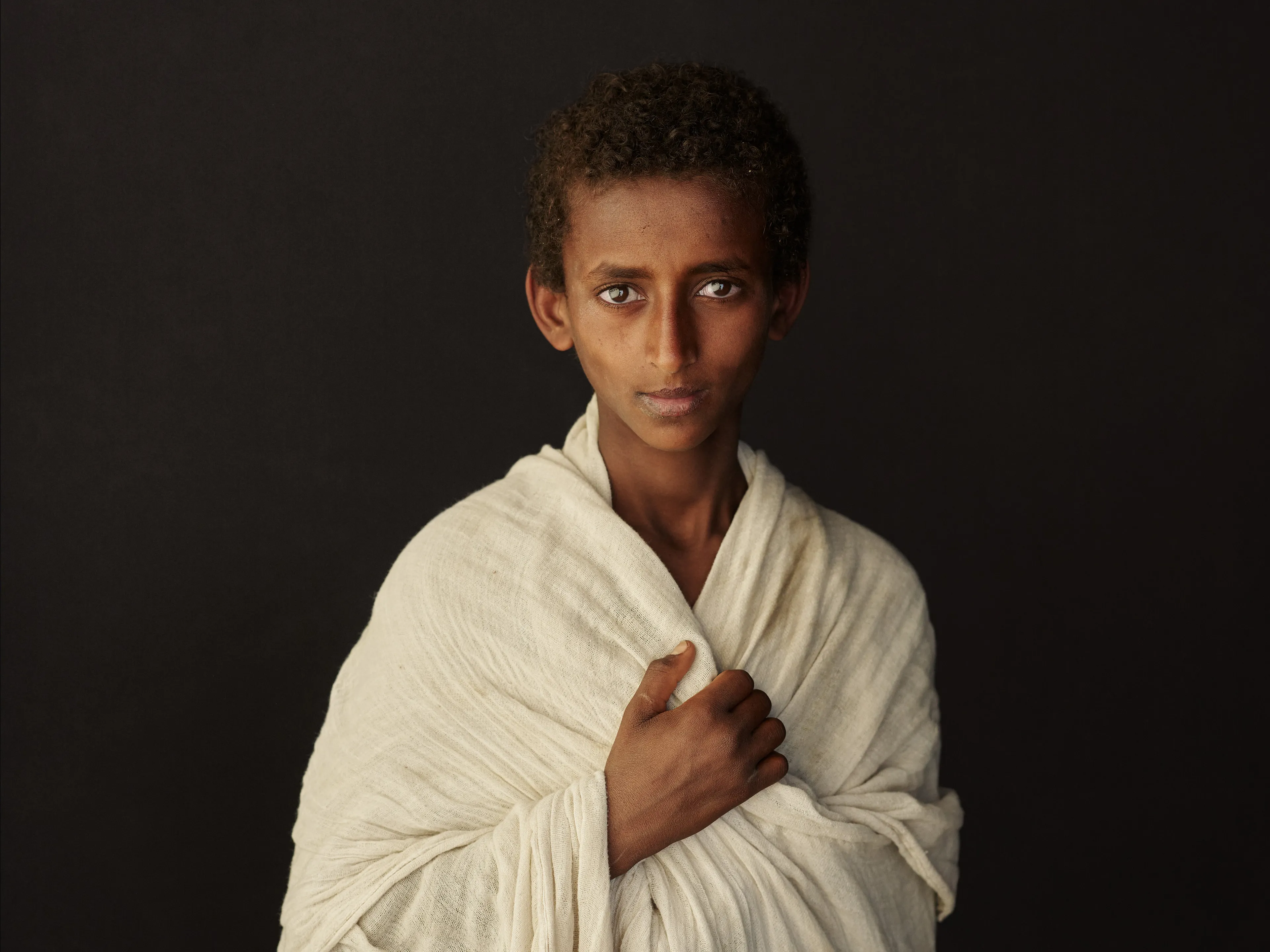 Joey L. returns to Quantum with Ethiopia - Portraits: a collection directly linked to his genesis drop during Season 1. For every environmental image from Joey’s first drop, there is a portrait companion.

“For years, we dragged our nomadic studio tent across almost every region of Ethiopia. In our tent, everyone was equal. We captured pure magic. Every kind of person entered and had their portrait taken. The tent was our little fantasy world. It was the Ethiopia that we dreamed of, even when the reality outside of it was sometimes stricken with searing pain. Each portrait was a tribute to a distinct region of the country. Disconnected cultural threads were woven together in a cohesive visual style in order to truly see Ethiopia— which itself is a modern nation state, but is also the sum of all the diverse people who inhabit it.”

Joey’s portraits alternating on red and black backdrops are the visual backbone of my forthcoming book “Ethiopia” (Insight Editions, January 2023), which compiles 13 years of work.

The tent itself is a metal frame modified with diffusion material made out of netela fabric, which is a hand-spun cotton perfected by Dorze craftspeople. Every portrait is created with natural light molded from the tent itself and photographed on location.

Lalibela, Amhara Region, Ethiopia 
January 2021