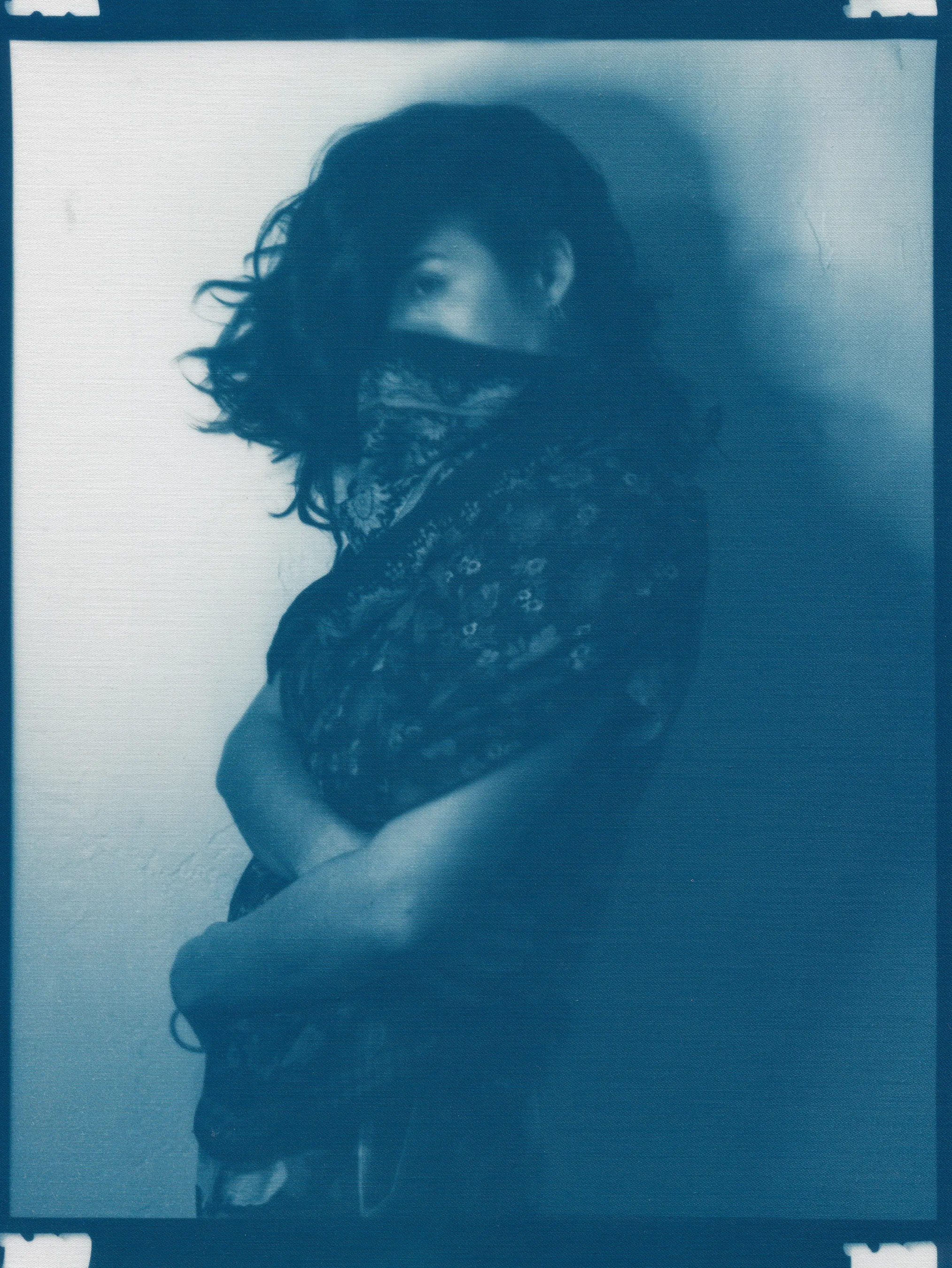 In the spring of 2020, panic surrounding COVID-19 erupted and mandatory shelter in place orders went into effect, forcing me to abandon long-planned portrait shoots, travel, and work in progress in my art studio.  I found myself in a 340 sq ft apartment in a Bay-area neighborhood that was emptying by the day.  The buzzing silence was profound. Two weeks into isolation, my drive to connect with others and make portraits only amplified.  With no safe way to do so, I spontaneously turned the camera on myself to create a portrait.  I printed it as a cyanotype, a simple nineteenth century photographic process that was only feasible due to the basic materials I happened to have on hand—a budget printer, transparency film and a package of fraying sheets of cotton pretreated with cyanotype chemicals.  At first, I exposed the prints in the unpredictable spring sunlight coming through my third floor apartment window.  As the seasons changed, I went on to make them in a garden, on the top of a car, or a patch of wobbly concrete tiles.  I rinsed them in water and varying shades of blue emerged.  I created a daily self-portrait using this technique for more than a year. 

I made most of these self-portraits inside various studio apartments that I lived in alone.  To make the earliest pictures in the series, I had to move a couch every day to create space against a white wall near a window.  I took others in fleeting spaces while traveling—in a guest room, in a medical examination room, during a pause in the wilderness, and later against the wall of an old California bungalow sandwiched between the mountains and the sea.  In the beginning, I often tried to cover as much of my body and face as possible as a commentary on my fear of the virus and my efforts to guard against it.  My armor and props ranged from common household items—potholders, tinfoil, dish towels, bedsheets, and toilet paper— to more telling evidence of the unusual consumption that resulted from being stuck indoors indefinitely— Amazon packaging, takeout bags and trash leftover from groceries purchased while wearing rubber gloves and sterilized in whatever way  was possible and later consumed.  These items shifted as the duration of the pandemic blurred into an unknown stretch of time. The portraits became less about those initial fears and more about confronting the boredom, anxiety, grief, and fatigue of living in indefinite isolation during a global pandemic. 

In total, I made these portraits for 377 consecutive days.  I made the first portrait on March 30th, 2020, a day when my head was raging in pain, my throat was constricted, and the fear that I might have contracted COVID hung over me before testing was easily accessible.  I made the last portrait on April 10th, 2021, the day I received my 2nd vaccination.  The days, weeks, and months in between feel like a fever dream—frayed and flickering in multitudes of blue.

2021_0306