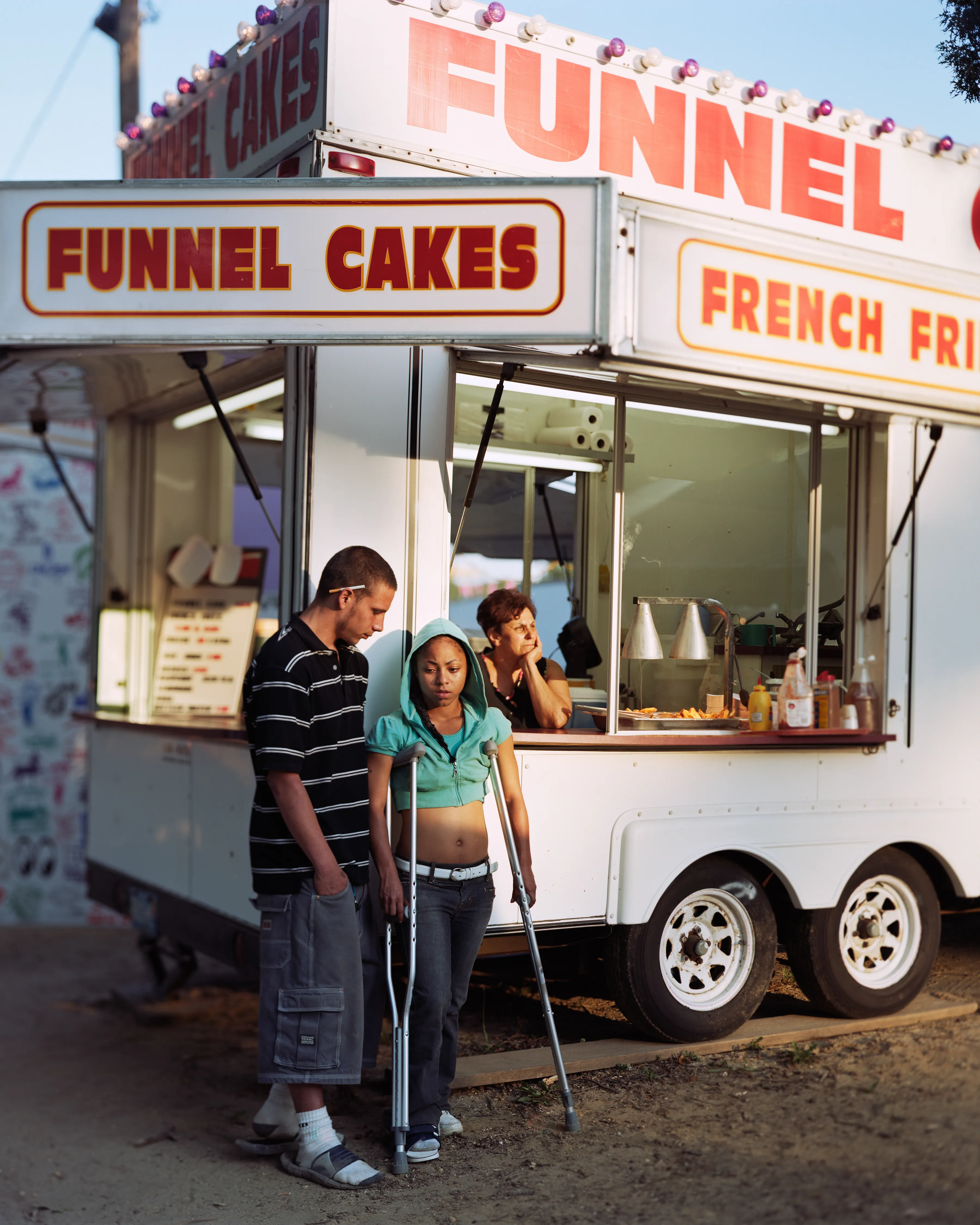 County fairs conjure ideas of tilt-a-whirls, stuffed animals, cotton candy, and blue ribbons. Rooted in local agricultural practices, fairs have been a ubiquitous part of American life for two centuries. In a reassuring way, they seem impervious to economic, technological, or fashion trends. For better or for worse, fairs look the same through the decades.

Though they are places to meet each other, there sure is a lot of loneliness at the fair. The series County Fair captures that duality: an adolescent boy looks up at the woman working the Hot Dog on a Stick stand as if she were a museum diorama; a little red haired girl haltingly befriends a robot; young couples, leaning and draped over each other, hide from the world in plain sight.


In my pictures, the dizzying atmosphere, colorfully decorated to provide amusement and distraction, is imbued with sadness and longing. These feelings, which permeate society, are laid bare at the fair. Sadness and longing circulate, intersect and incubate in the confined space between the ticket booths and the craft barns. People come out to participate and show, to see and be seen. Ultimately, connection remains elusive. 


