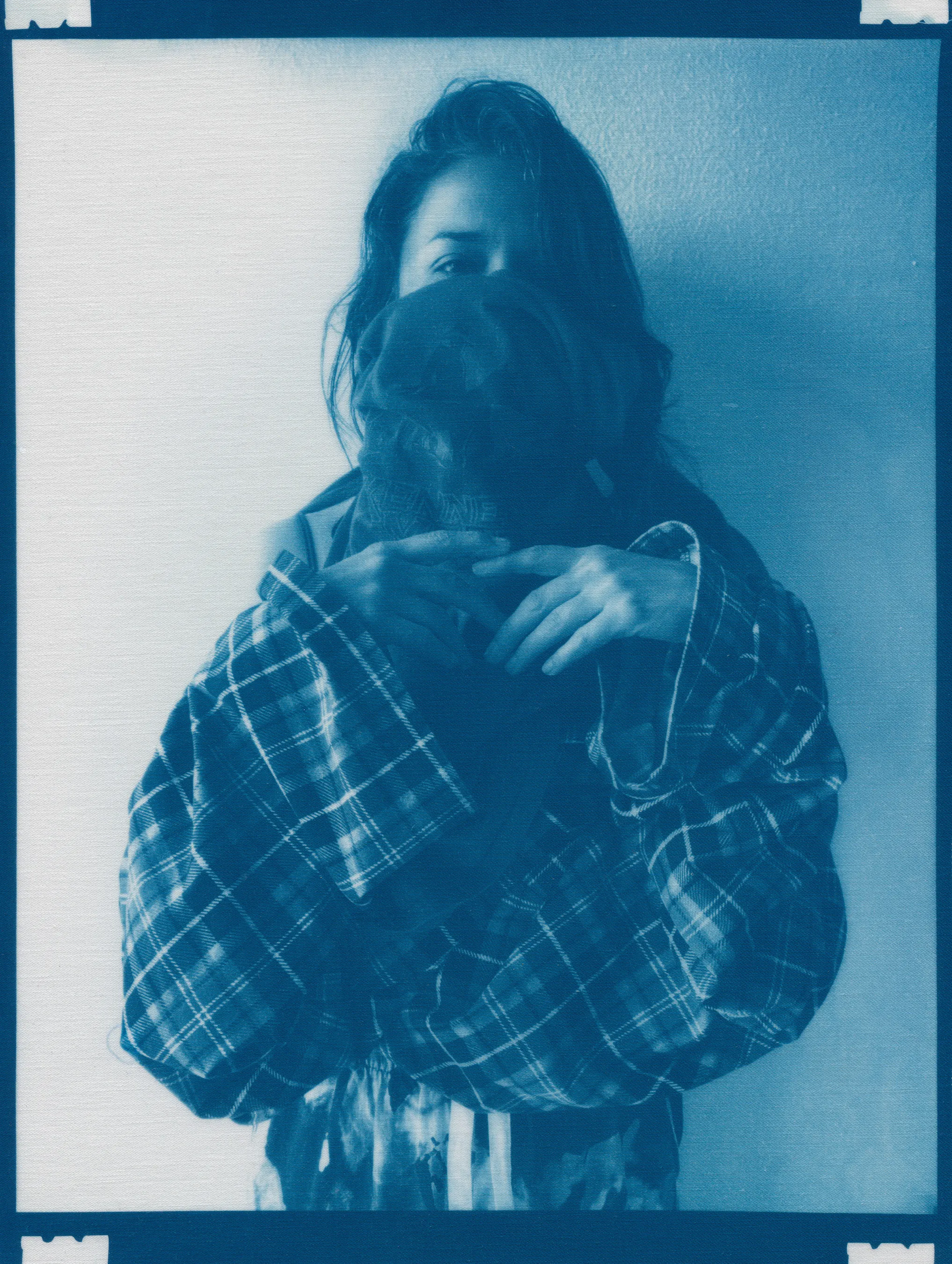 In the spring of 2020, panic surrounding COVID-19 erupted and mandatory shelter in place orders went into effect, forcing me to abandon long-planned portrait shoots, travel, and work in progress in my art studio.  I found myself in a 340 sq ft apartment in a Bay-area neighborhood that was emptying by the day.  The buzzing silence was profound. Two weeks into isolation, my drive to connect with others and make portraits only amplified.  With no safe way to do so, I spontaneously turned the camera on myself to create a portrait.  I printed it as a cyanotype, a simple nineteenth century photographic process that was only feasible due to the basic materials I happened to have on hand—a budget printer, transparency film and a package of fraying sheets of cotton pretreated with cyanotype chemicals.  At first, I exposed the prints in the unpredictable spring sunlight coming through my third floor apartment window.  As the seasons changed, I went on to make them in a garden, on the top of a car, or a patch of wobbly concrete tiles.  I rinsed them in water and varying shades of blue emerged.  I created a daily self-portrait using this technique for more than a year. 

I made most of these self-portraits inside various studio apartments that I lived in alone.  To make the earliest pictures in the series, I had to move a couch every day to create space against a white wall near a window.  I took others in fleeting spaces while traveling—in a guest room, in a medical examination room, during a pause in the wilderness, and later against the wall of an old California bungalow sandwiched between the mountains and the sea.  In the beginning, I often tried to cover as much of my body and face as possible as a commentary on my fear of the virus and my efforts to guard against it.  My armor and props ranged from common household items—potholders, tinfoil, dish towels, bedsheets, and toilet paper— to more telling evidence of the unusual consumption that resulted from being stuck indoors indefinitely— Amazon packaging, takeout bags and trash leftover from groceries purchased while wearing rubber gloves and sterilized in whatever way  was possible and later consumed.  These items shifted as the duration of the pandemic blurred into an unknown stretch of time. The portraits became less about those initial fears and more about confronting the boredom, anxiety, grief, and fatigue of living in indefinite isolation during a global pandemic. 

In total, I made these portraits for 377 consecutive days.  I made the first portrait on March 30th, 2020, a day when my head was raging in pain, my throat was constricted, and the fear that I might have contracted COVID hung over me before testing was easily accessible.  I made the last portrait on April 10th, 2021, the day I received my 2nd vaccination.  The days, weeks, and months in between feel like a fever dream—frayed and flickering in multitudes of blue.

2021_0320