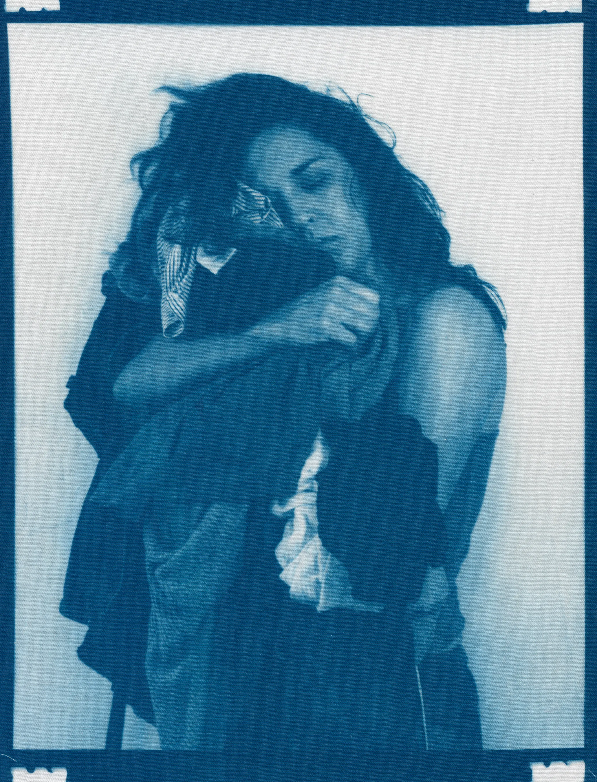 In the spring of 2020, panic surrounding COVID-19 erupted and mandatory shelter in place orders went into effect, forcing me to abandon long-planned portrait shoots, travel, and work in progress in my art studio.  I found myself in a 340 sq ft apartment in a Bay-area neighborhood that was emptying by the day.  The buzzing silence was profound. Two weeks into isolation, my drive to connect with others and make portraits only amplified.  With no safe way to do so, I spontaneously turned the camera on myself to create a portrait.  I printed it as a cyanotype, a simple nineteenth century photographic process that was only feasible due to the basic materials I happened to have on hand—a budget printer, transparency film and a package of fraying sheets of cotton pretreated with cyanotype chemicals.  At first, I exposed the prints in the unpredictable spring sunlight coming through my third floor apartment window.  As the seasons changed, I went on to make them in a garden, on the top of a car, or a patch of wobbly concrete tiles.  I rinsed them in water and varying shades of blue emerged.  I created a daily self-portrait using this technique for more than a year. 

I made most of these self-portraits inside various studio apartments that I lived in alone.  To make the earliest pictures in the series, I had to move a couch every day to create space against a white wall near a window.  I took others in fleeting spaces while traveling—in a guest room, in a medical examination room, during a pause in the wilderness, and later against the wall of an old California bungalow sandwiched between the mountains and the sea.  In the beginning, I often tried to cover as much of my body and face as possible as a commentary on my fear of the virus and my efforts to guard against it.  My armor and props ranged from common household items—potholders, tinfoil, dish towels, bedsheets, and toilet paper— to more telling evidence of the unusual consumption that resulted from being stuck indoors indefinitely— Amazon packaging, takeout bags and trash leftover from groceries purchased while wearing rubber gloves and sterilized in whatever way  was possible and later consumed.  These items shifted as the duration of the pandemic blurred into an unknown stretch of time. The portraits became less about those initial fears and more about confronting the boredom, anxiety, grief, and fatigue of living in indefinite isolation during a global pandemic. 

In total, I made these portraits for 377 consecutive days.  I made the first portrait on March 30th, 2020, a day when my head was raging in pain, my throat was constricted, and the fear that I might have contracted COVID hung over me before testing was easily accessible.  I made the last portrait on April 10th, 2021, the day I received my 2nd vaccination.  The days, weeks, and months in between feel like a fever dream—frayed and flickering in multitudes of blue.

2021_0403
