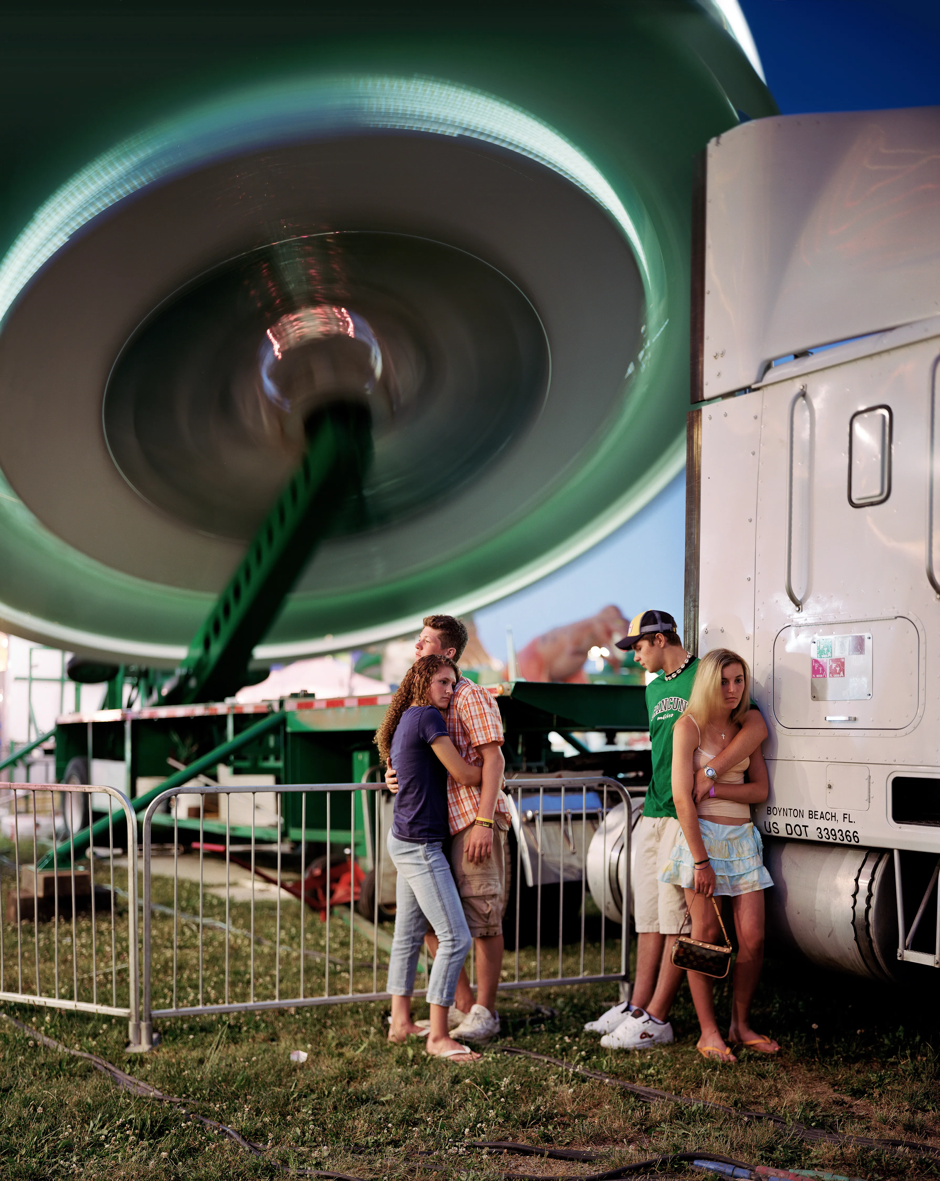 County fairs conjure ideas of tilt-a-whirls, stuffed animals, cotton candy, and blue ribbons. Rooted in local agricultural practices, fairs have been a ubiquitous part of American life for two centuries. In a reassuring way, they seem impervious to economic, technological, or fashion trends. For better or for worse, fairs look the same through the decades.

Though they are places to meet each other, there sure is a lot of loneliness at the fair. The series County Fair captures that duality: an adolescent boy looks up at the woman working the Hot Dog on a Stick stand as if she were a museum diorama; a little red haired girl haltingly befriends a robot; young couples, leaning and draped over each other, hide from the world in plain sight.


In my pictures, the dizzying atmosphere, colorfully decorated to provide amusement and distraction, is imbued with sadness and longing. These feelings, which permeate society, are laid bare at the fair. Sadness and longing circulate, intersect and incubate in the confined space between the ticket booths and the craft barns. People come out to participate and show, to see and be seen. Ultimately, connection remains elusive. 

