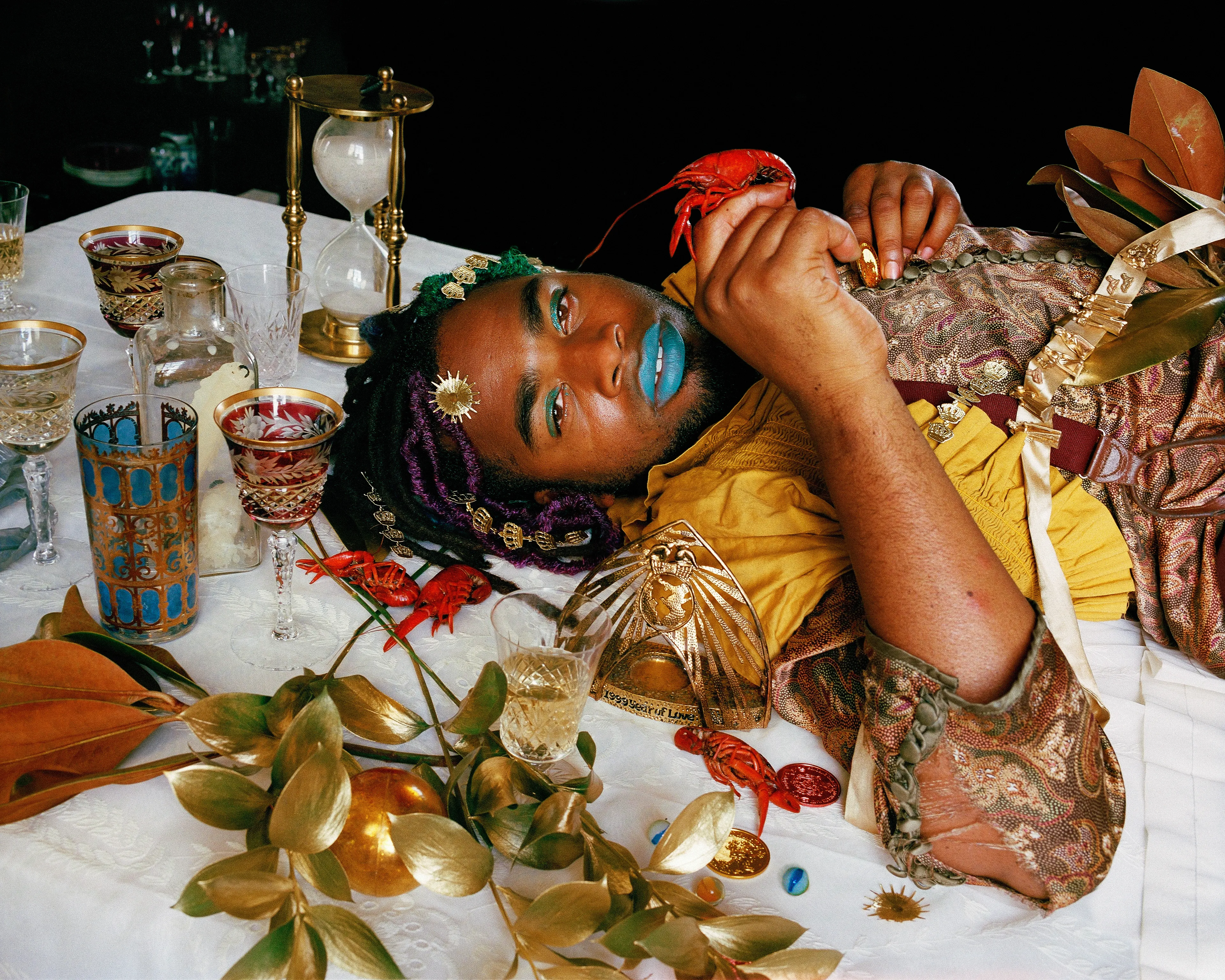 Kristina E. Knipe's Talisman offer details in excess--a rhinestone shimmering behind the wing of a dead cockroach, a cracked magnifying glass amplifies a passionflower, oxidation on a finger from a brass ring turns. Transgressing the historic photographic ordering of society, she embraces mess and its attendant spectacle. Queer ritual disrupts engrained hierarchies, rejecting outmoded classifications. In New Orleans the altars, masquerades, and decadences create a visual and material excess that heals as it depletes. She adorns her sitters in shared materials to create a transformative experience of pleasure.

My roommate jumped out of a building on Mardi Gras day while rolling on ecstasy. She broke her ankle and spent that night in a hospital bed. I couldn't be of any help as I had scratched my cornea with glitter. On Easter, a mended friend gave us a broken wheelchair to roll around the French Quarter chasing after Ostara and the Gay Easter Parade. To memorialize her injured ankle we arranged a tableau of broken goblets, molding banana blossoms, her old cast, and opulent detritus. Debauchery scars the body.

These 4x5 negatives offer details in excess--a rhinestone shimmering behind the wing of a dead cockroach, a cracked magnifying glass amplifies a passionflower, oxidation from a brass ring turns Antony’s finger green. To transgress the historic photographic ordering of society, I embrace mess and its attendant spectacle. Queer ritual disrupts engrained hierarchies, rejecting outmoded classifications. In New Orleans the altars, masquerades, and decadences create a visual and material excess that heals as it depletes. I adorn my sitters in our shared materials to create a transformative experience of pleasure.

The concrete reality of those I photograph consists of the burden of maintaining mental health without healthcare, of being a person whose role is to make other’s spaces beautiful which by necessity means repurposing objects for their own, of having traumatic experiences with no recourse, cycles of depression in the service of the hospitality industry, expressing gender and sexuality despite judgment, and persisting in generative work through loss. My story is intertwined with those I photograph- we cohabit spaces and experiences within a community of artists, activists, and healers.
