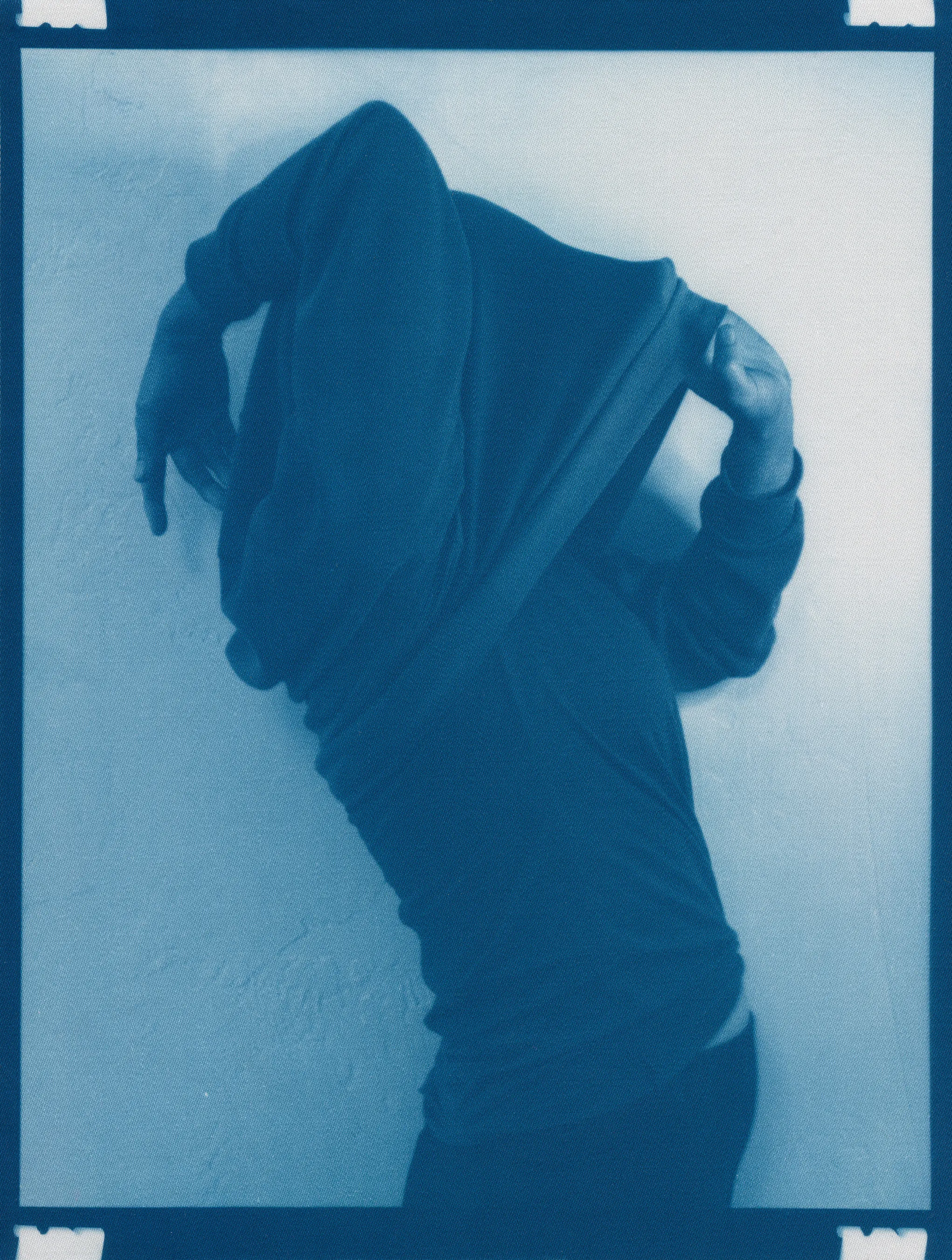In the spring of 2020, panic surrounding COVID-19 erupted and mandatory shelter in place orders went into effect, forcing me to abandon long-planned portrait shoots, travel, and work in progress in my art studio.  I found myself in a 340 sq ft apartment in a Bay-area neighborhood that was emptying by the day.  The buzzing silence was profound. Two weeks into isolation, my drive to connect with others and make portraits only amplified.  With no safe way to do so, I spontaneously turned the camera on myself to create a portrait.  I printed it as a cyanotype, a simple nineteenth century photographic process that was only feasible due to the basic materials I happened to have on hand—a budget printer, transparency film and a package of fraying sheets of cotton pretreated with cyanotype chemicals.  At first, I exposed the prints in the unpredictable spring sunlight coming through my third floor apartment window.  As the seasons changed, I went on to make them in a garden, on the top of a car, or a patch of wobbly concrete tiles.  I rinsed them in water and varying shades of blue emerged.  I created a daily self-portrait using this technique for more than a year. 

I made most of these self-portraits inside various studio apartments that I lived in alone.  To make the earliest pictures in the series, I had to move a couch every day to create space against a white wall near a window.  I took others in fleeting spaces while traveling—in a guest room, in a medical examination room, during a pause in the wilderness, and later against the wall of an old California bungalow sandwiched between the mountains and the sea.  In the beginning, I often tried to cover as much of my body and face as possible as a commentary on my fear of the virus and my efforts to guard against it.  My armor and props ranged from common household items—potholders, tinfoil, dish towels, bedsheets, and toilet paper— to more telling evidence of the unusual consumption that resulted from being stuck indoors indefinitely— Amazon packaging, takeout bags and trash leftover from groceries purchased while wearing rubber gloves and sterilized in whatever way  was possible and later consumed.  These items shifted as the duration of the pandemic blurred into an unknown stretch of time. The portraits became less about those initial fears and more about confronting the boredom, anxiety, grief, and fatigue of living in indefinite isolation during a global pandemic. 

In total, I made these portraits for 377 consecutive days.  I made the first portrait on March 30th, 2020, a day when my head was raging in pain, my throat was constricted, and the fear that I might have contracted COVID hung over me before testing was easily accessible.  I made the last portrait on April 10th, 2021, the day I received my 2nd vaccination.  The days, weeks, and months in between feel like a fever dream—frayed and flickering in multitudes of blue.

2021_0406