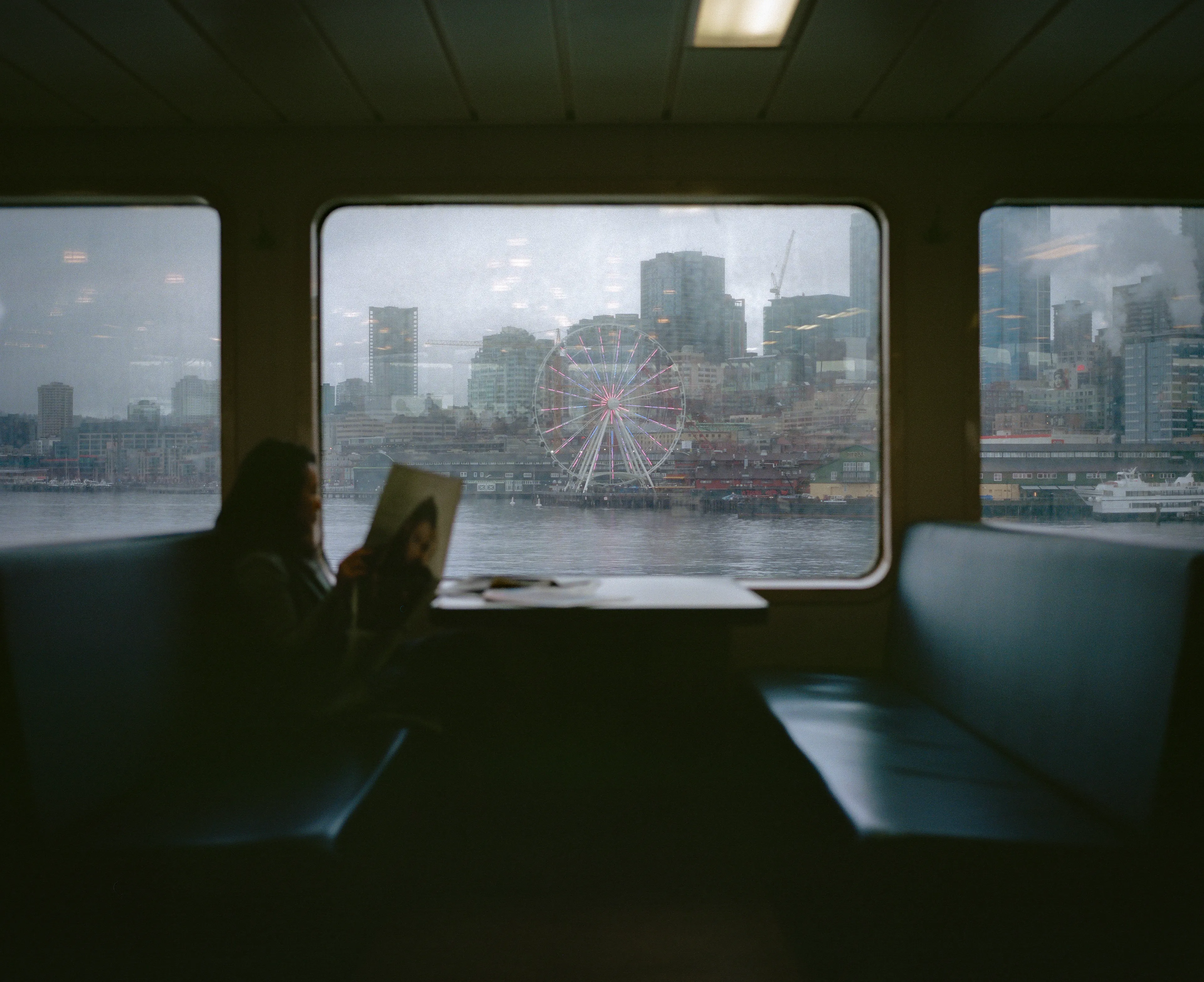 A lone passenger sat by the window, engrossed in the morning newspaper. The faint outline of the iconic Seattle Great Wheel and the city's skyline framed in the misty glass to their left offered a stark contrast to the somber weather outside, providing a serene backdrop to their quiet reading moment.