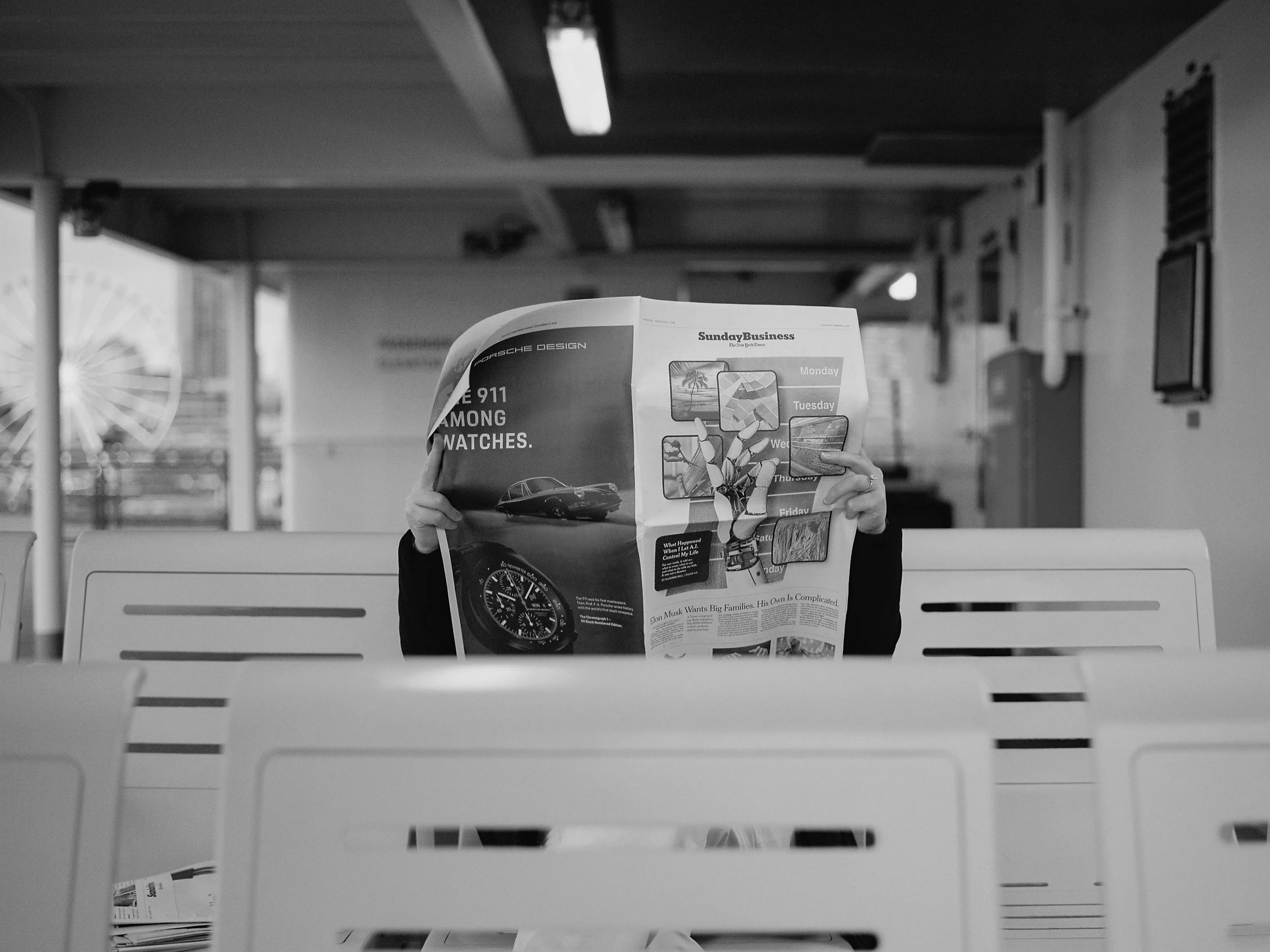 I had AI describe this photograph:

"This photograph shows a person seated on a bench, holding up a newspaper that obscures their face. The setting appears to be an indoor waiting area with rows of white seats. In the background, there is a blurred view of a ferris wheel and some architectural elements, suggesting the location might be near an amusement park or waterfront. The overall tone of the image is casual and candid, with a soft focus on the background and sharp details on the newspaper. The lighting appears natural, coming from windows or an open area behind the photographer."