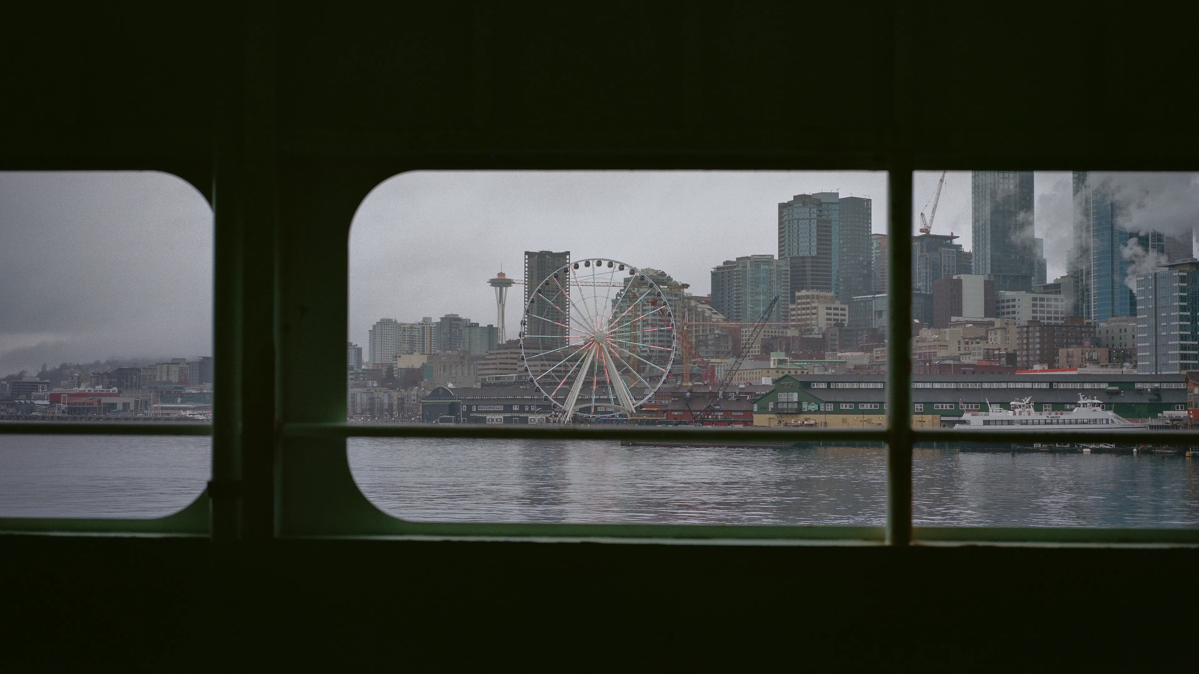 Leaving Seattle on a cold winter morning via ferry, the distant Ferris wheel stood silent against the gray sky, a symbol of the city's vibrant waterfront. As the ferry calmly glided away, the wheel's colorful spokes faded into the misty horizon.