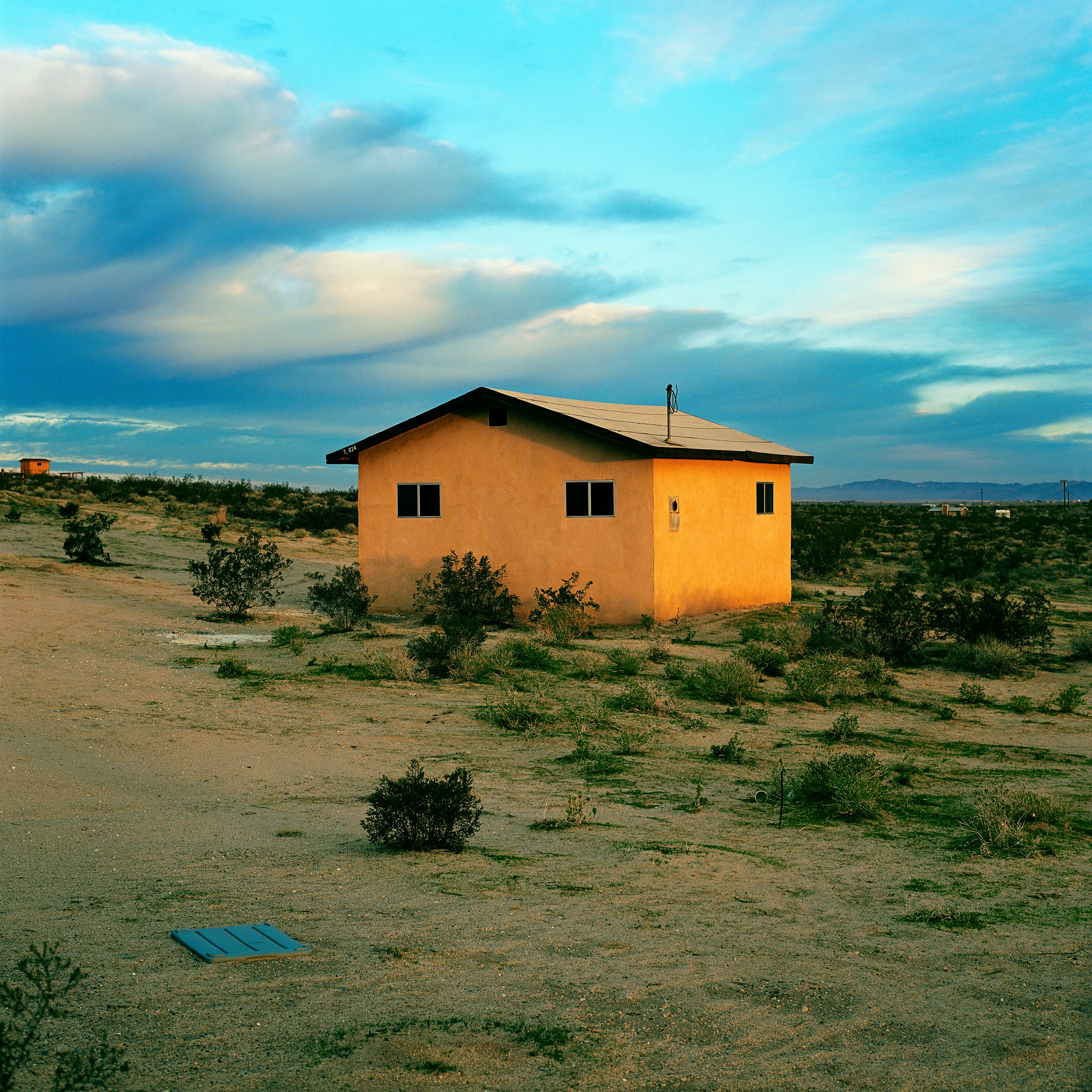 “These are photographs of isolated structures in the high desert of Southern California. All of the images are made in the east end of the Morongo Valley Basin, Wonder Valley, and the area surrounding the town of Twenty-Nine Palms. This is an area approximately one hundred- and fifty-miles due East of Los Angeles. From the oceans edge in L.A., to the area that I am photographing, the landscape is continuously occupied. This area is the final edge of a vast urban expanse. For social, financial, or personal reasons individuals are pushed, or pulled, or simply seek this literal cultural edge. Beyond this point is unoccupied desert.  In the past I have perused projects which dealt with the iconography of transcendence and the sublime. In other projects I have investigated the literal desire to get "outside" of or beyond the cultural (a desire with the ironic consequence of transforming the natural into the very condition one wishes to escape). It is this conceptual trajectory which has led me to this current body of work. This current body of work is an extension of the investigation begun in the "four landscape" portfolio. The structural rigor of that work has been replaced with concerns which are aesthetic and social. On the simplest level I am drawn to the vernacular character of the architecture (painted with a Home Depot palette) as well as the amazing visual character of these structures on this vast desert plain lit by an extraordinary light. However, at base, my primary interest is to create images which are iconographic of a desire. A desire to be "beyond," a desire to be alone, a sign of man on the landscape”. - John Divola.

Edition: 1 of 1