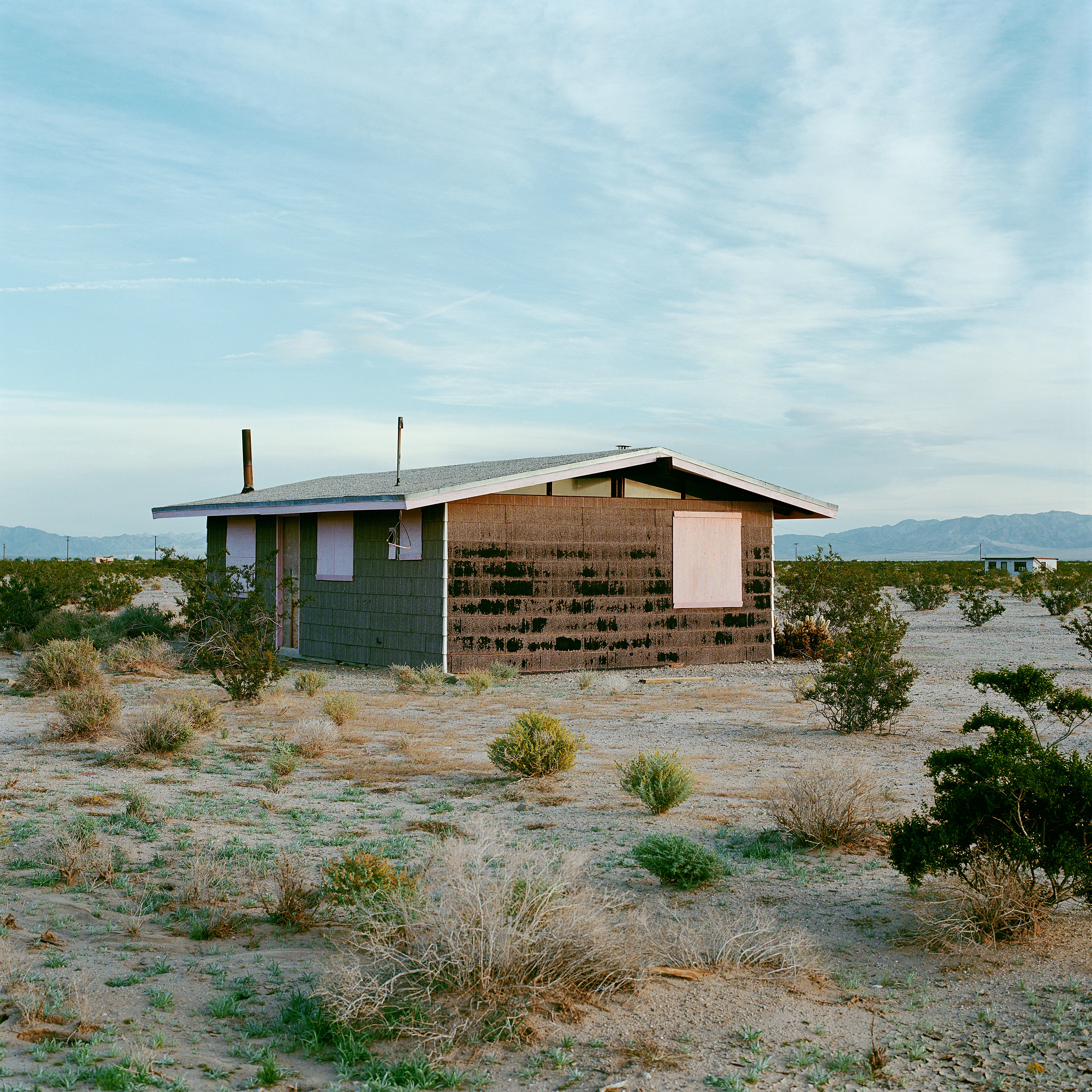 “These are photographs of isolated structures in the high desert of Southern California. All of the images are made in the east end of the Morongo Valley Basin, Wonder Valley, and the area surrounding the town of Twenty-Nine Palms. This is an area approximately one hundred- and fifty-miles due East of Los Angeles. From the oceans edge in L.A., to the area that I am photographing, the landscape is continuously occupied. This area is the final edge of a vast urban expanse. For social, financial, or personal reasons individuals are pushed, or pulled, or simply seek this literal cultural edge. Beyond this point is unoccupied desert.  In the past I have perused projects which dealt with the iconography of transcendence and the sublime. In other projects I have investigated the literal desire to get "outside" of or beyond the cultural (a desire with the ironic consequence of transforming the natural into the very condition one wishes to escape). It is this conceptual trajectory which has led me to this current body of work. This current body of work is an extension of the investigation begun in the "four landscape" portfolio. The structural rigor of that work has been replaced with concerns which are aesthetic and social. On the simplest level I am drawn to the vernacular character of the architecture (painted with a Home Depot palette) as well as the amazing visual character of these structures on this vast desert plain lit by an extraordinary light. However, at base, my primary interest is to create images which are iconographic of a desire. A desire to be "beyond", a desire to be alone, a sign of man on the landscape”. - John Divola.

Edition: 1 of 1