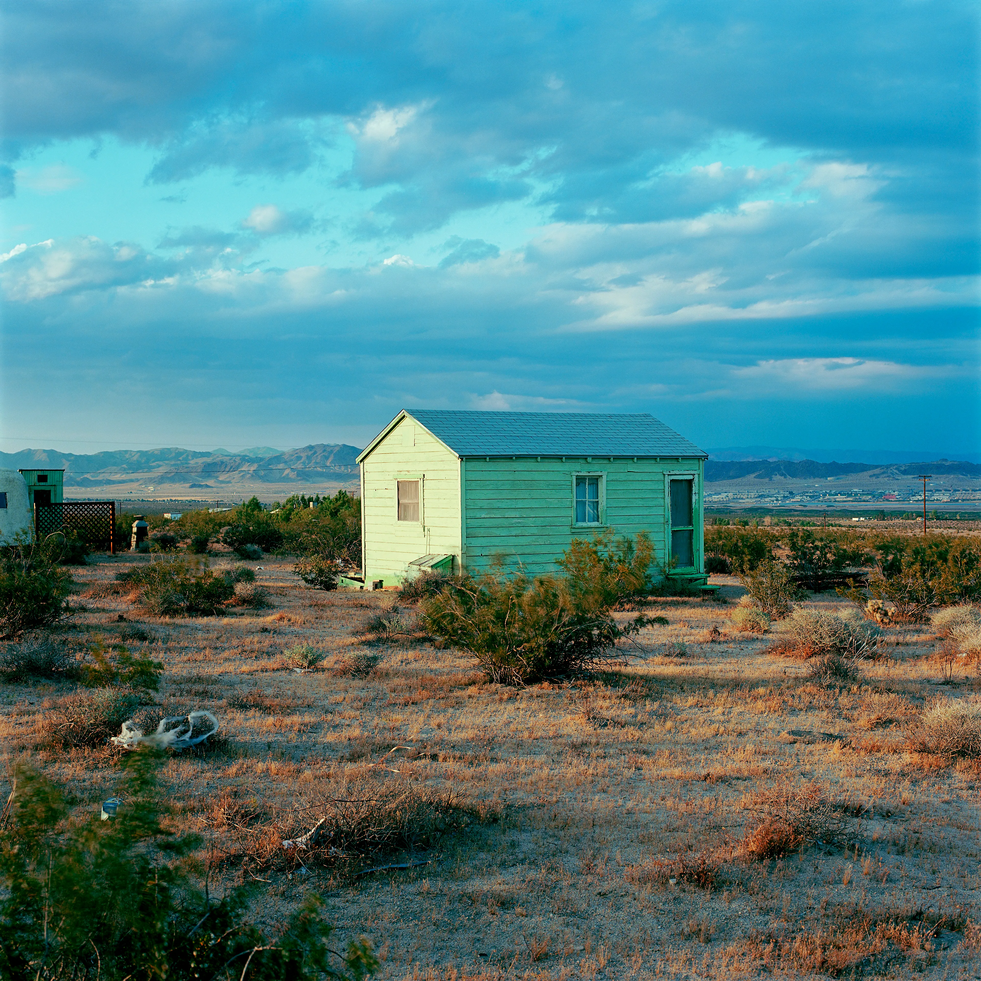 “These are photographs of isolated structures in the high desert of Southern California. All of the images are made in the east end of the Morongo Valley Basin, Wonder Valley, and the area surrounding the town of Twenty-Nine Palms. This is an area approximately one hundred- and fifty-miles due East of Los Angeles. From the oceans edge in L.A., to the area that I am photographing, the landscape is continuously occupied. This area is the final edge of a vast urban expanse. For social, financial, or personal reasons individuals are pushed, or pulled, or simply seek this literal cultural edge. Beyond this point is unoccupied desert.  In the past I have perused projects which dealt with the iconography of transcendence and the sublime. In other projects I have investigated the literal desire to get "outside" of or beyond the cultural (a desire with the ironic consequence of transforming the natural into the very condition one wishes to escape). It is this conceptual trajectory which has led me to this current body of work. This current body of work is an extension of the investigation begun in the "four landscape" portfolio. The structural rigor of that work has been replaced with concerns which are aesthetic and social. On the simplest level I am drawn to the vernacular character of the architecture (painted with a Home Depot palette) as well as the amazing visual character of these structures on this vast desert plain lit by an extraordinary light. However, at base, my primary interest is to create images which are iconographic of a desire. A desire to be "beyond", a desire to be alone, a sign of man on the landscape”. - John Divola.

Edition: 1 of 1