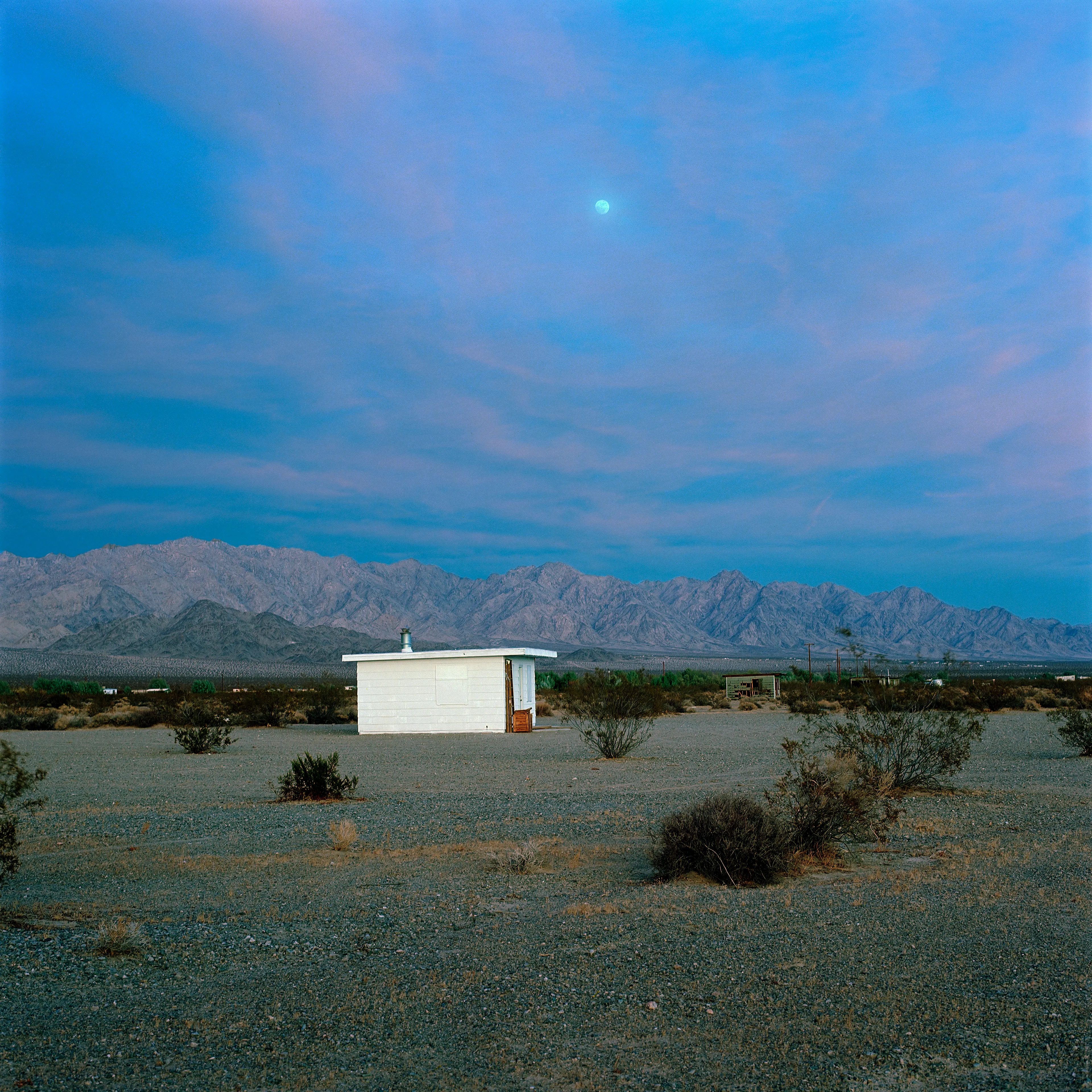 “These are photographs of isolated structures in the high desert of Southern California. All of the images are made in the east end of the Morongo Valley Basin, Wonder Valley, and the area surrounding the town of Twenty-Nine Palms. This is an area approximately one hundred- and fifty-miles due East of Los Angeles. From the oceans edge in L.A., to the area that I am photographing, the landscape is continuously occupied. This area is the final edge of a vast urban expanse. For social, financial, or personal reasons individuals are pushed, or pulled, or simply seek this literal cultural edge. Beyond this point is unoccupied desert.  In the past I have perused projects which dealt with the iconography of transcendence and the sublime. In other projects I have investigated the literal desire to get "outside" of or beyond the cultural (a desire with the ironic consequence of transforming the natural into the very condition one wishes to escape). It is this conceptual trajectory which has led me to this current body of work. This current body of work is an extension of the investigation begun in the "four landscape" portfolio." The structural rigor of that work has been replaced with concerns which are aesthetic and social. On the simplest level I am drawn to the vernacular character of the architecture (painted with a Home Depot palette) as well as the amazing visual character of these structures on this vast desert plain lit by an extraordinary light. However, at base, my primary interest is to create images which are iconographic of a desire. A desire to be "beyond," a desire to be alone, a sign of man on the landscape”. - John Divola.

Edition: 1 of 1