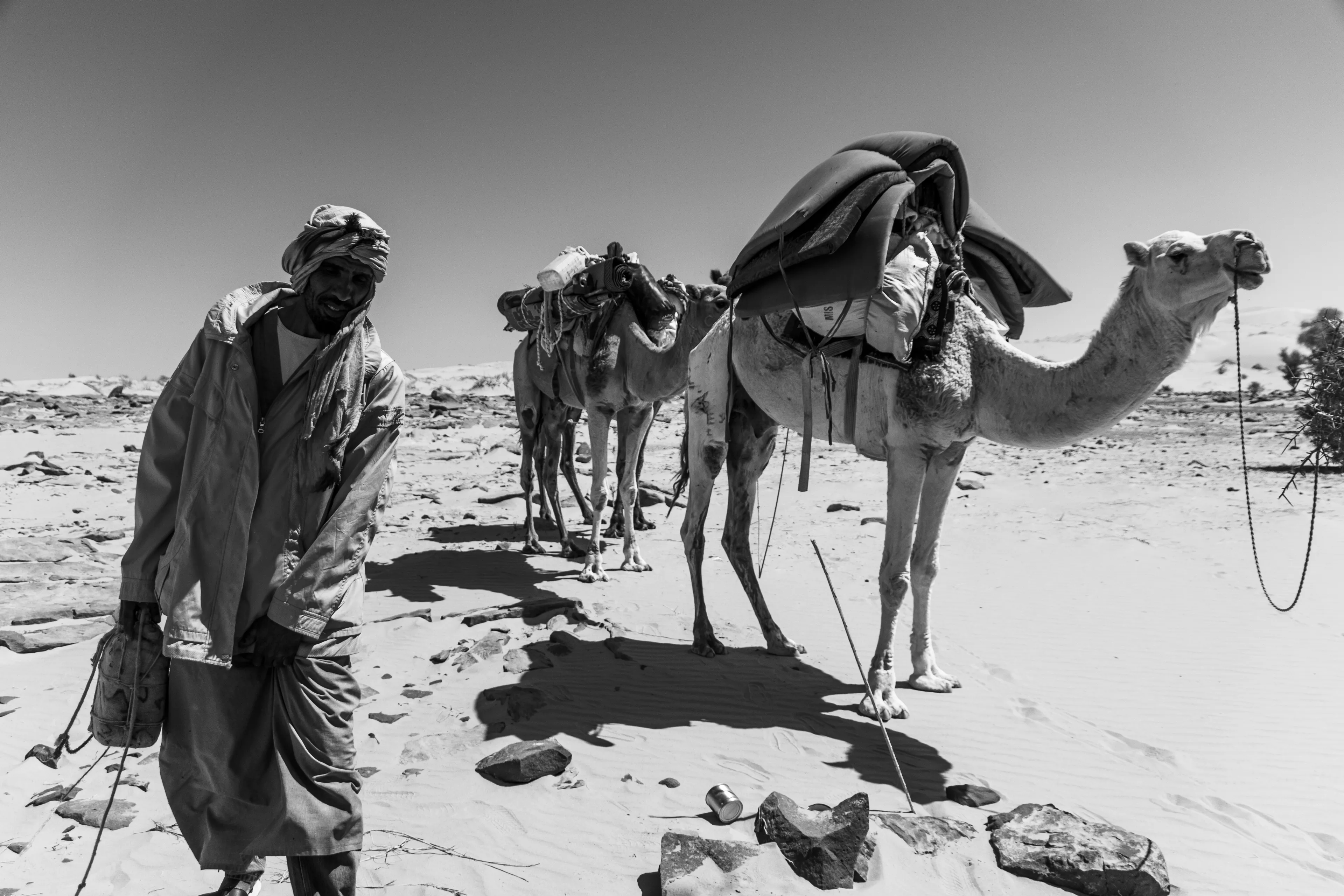 En route across the desert, we let the camels recharge at a lone bush, their long necks reaching for its sparse leaves. Beside them, the chamelier bends forward, his posture echoing theirs, as if man and animal were shaped by the same rhythm.