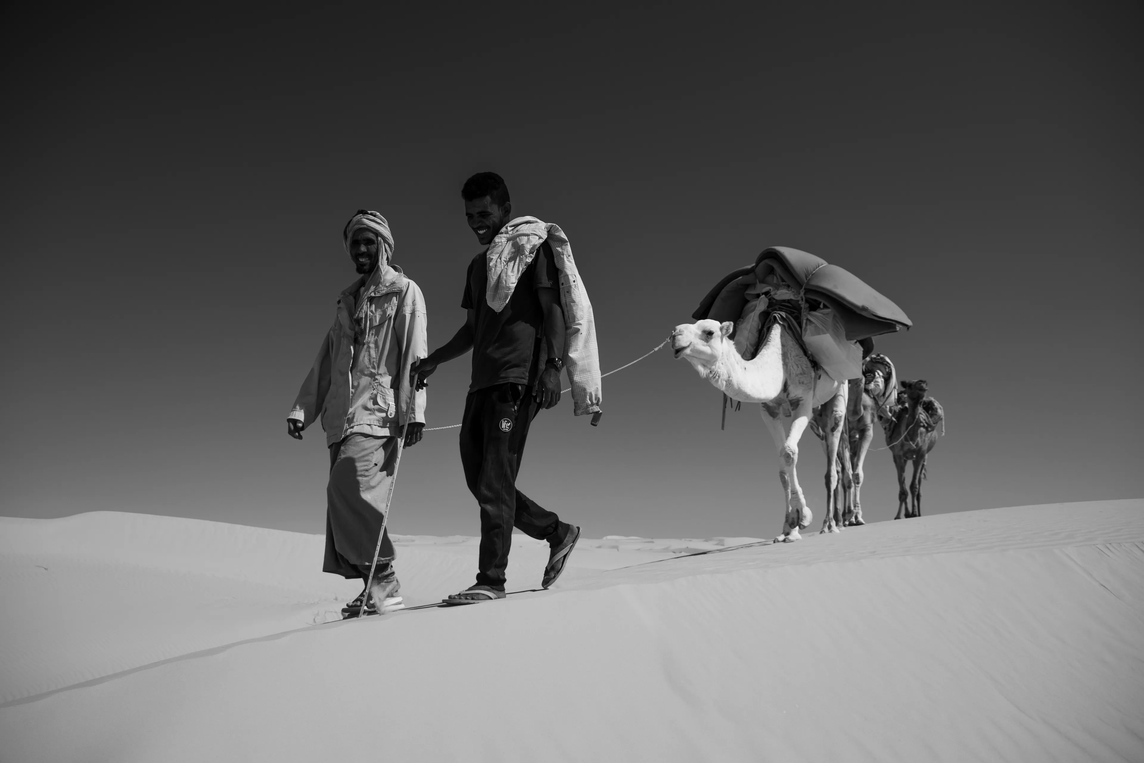 Pacing steadily across the sands, our two chameliers smile as they lead our camel caravan, carrying supplies from Chinguetti toward the Oasis de La Gueila.