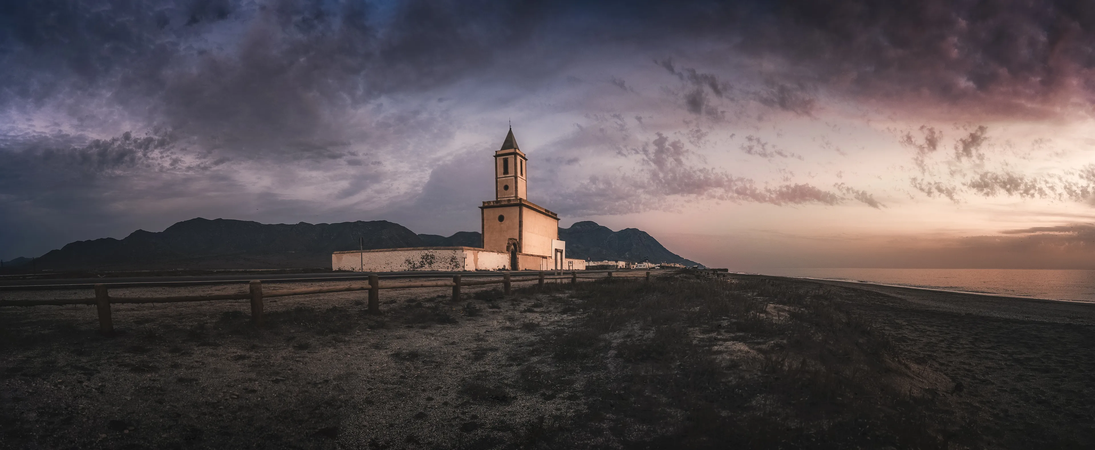 It was a late autumn afternoon, the kind where the sun hurries toward the horizon, leaving the sky streaked with shifting colors and long, soft shadows. I hadn’t planned to come to the Church of Las Salinas. It was a spur-of-the-moment decision, a quiet escape from the noise of everyday life. I parked a short distance away and walked slowly, feeling the rough texture of the ground under my feet and listening to the faint crash of the waves in the distance.

The church stood there, quiet and timeless, its worn facade telling stories of another era. Once, its walls must have held the echoes of prayers and songs, but now silence had taken over. It wasn’t an empty silence, though—it was a welcoming one, as if the place invited you to stop, to take a moment, and to listen with more than just your ears.

I sat down at the edge of the wooden path, facing the church, letting the cool evening breeze wash over me. My camera rested in my hands, but I wasn’t in a hurry to shoot. I wanted more than just a photo. I wanted to capture what I felt: the vastness of the sky, brushed with soft, fading light, and the quiet weight of the mountains in the distance, standing like guardians of this forgotten corner.

As I adjusted the lens, an elderly man walked by, leaning on his cane. He stopped when he saw me, offering a kind smile.  
“Beautiful place, isn’t it?” he said, his voice steady and warm.  
I nodded.  
“This used to be the heart of the salinas,” he continued, gesturing toward the church. “The workers would gather here after their shifts, grateful for what this land gave them. It’s been years now… but it still holds its magic, don’t you think?”

I nodded again, wanting to say more but unsure of what words could match the moment. With a small tilt of his head, he turned and continued on his way toward the beach, leaving me with his words—and a strange, bittersweet feeling.

As the last of the sunlight faded, leaving a deep purple glow in the sky, I took a few final moments to breathe it all in. The crisp air, the sound of the distant waves, the quiet presence of the church—it all felt like more than a scene. It felt alive, a piece of history lingering in the present.

When I finally drove away, I glanced back through the rearview mirror. The church was still there, its silhouette standing firm against the growing night. I couldn’t help but smile. Some places stay with you long after you leave, and I knew this was one of them. I would come back, because places like this don’t just call to you once. They become a part of you.