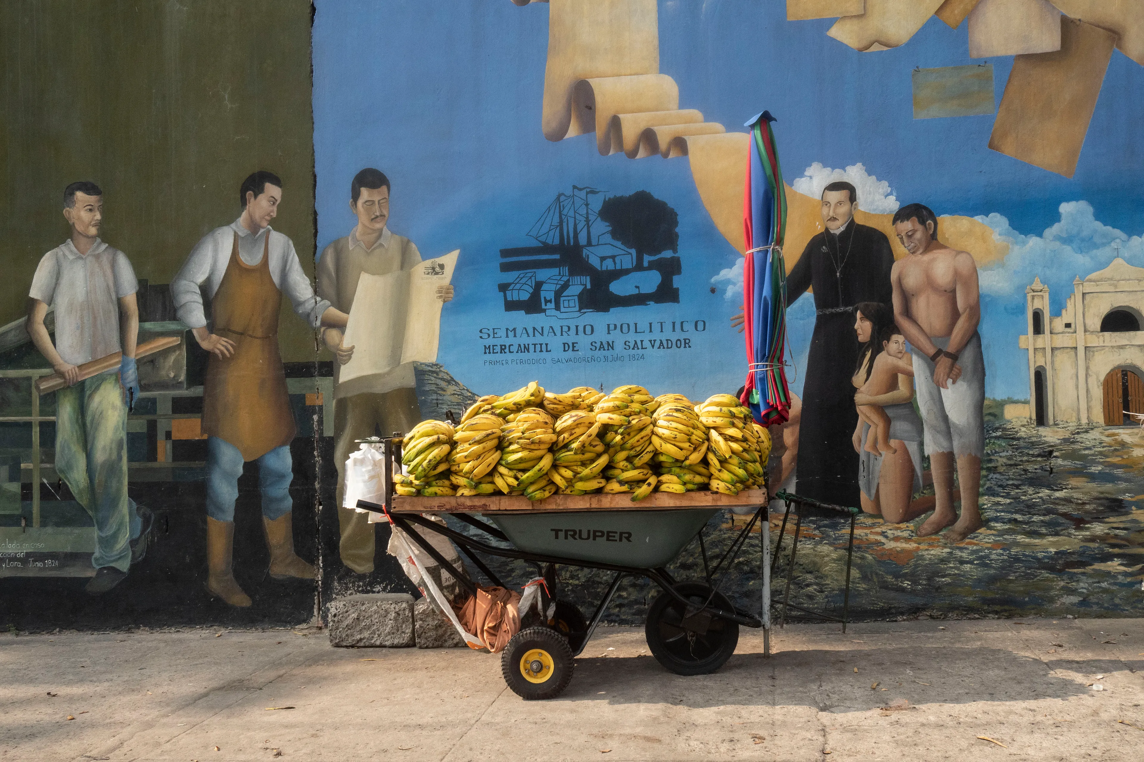 « Bitcoin Nation » #33 by Thomas Dworzak

EL SALVADOR, San Salvador, 2022/3-4. History of EL SALVADOR mural. Banana seller. Neighbouring Honduras was the original "Banana Republic".

« Bitcoin Nation » - El Salvador - Thomas Dworzak / Magnum Photos

Many photographers and journalists of my generation, interested in war and conflict, would at one point read Richard Kapusciznsky's all-time classic "The Soccer War" and watch Oliver Stone's "Salvador. » The latternow feels insanely cheesy. With the country's troubled history in mind and never having been to Central America, I found "Bitcoin Nation - El Salvador" a challenging theme to explore for this NFT project. 

I set out to straightforwardly document this alien and invisible world of bitcoin, and being 'in the realm of crypto,' I hoped it would give me access to President Nayib Bukele's surprise project. Last year he had suddenly declared Bitcoin the official currency of El Salvador - which until now was only the US dollar. The 40-year-old president of Palestinian origin started out as an anti-establishment candidate, describing himself as "the world's coolest dictator," but soon took an authoritarian turn. Yet he continues drumming the bitcoin beat, and bitcoin celebrities from all around the world keep flocking to El Salvador.
The Pacific beach town of El Zonte became "Bitcoin Beach," and "Volcano Bonds" and "Bitcoin City" are meant to be the future.

When official doors remained closed for me, I started digging for clues in the county's troubled past and present or anything, even remotely, related to the bitcoin world. Vignettes of the bloody civil war regularly popped up, and I came across a few "bitcoin events." The sudden spike of almost a hundred homicides in the course of one weekend in late March, due to what seemed a breakdown of a secret deal between the government and the two main gangs, had President Bukele declare a state of emergency, martial law and intensify the war on the gangs. I could see fringes of it. But nothing compared to the brutality of
the spectacularly filmed daily government propaganda videos of mass arrests and more-than-rough prisoner handlings I watched on TV, Twitter, and Instagram.

It was at a small bitcoin event that I found a hint why I may have been shunned and didn't get any access to show the "shiny world of Bitcoin Nation," as I had hoped. My NFT ticket was definitely the "wrong church" in the Bitcoin world. To paraphrase one of the speakers: "If I want to gamble, I go to Vegas and party with beautiful hookers, instead of being a loser, sitting around in my basement, frustrated, minting NFTs. Shitcoins."

El Salvador and the President were meant to be guests of honor at the Bitcoin 2022 conference in Miami. But due to the internal tension, he canceled the visit, and the 30+ thousand delegates had to discuss the future of bitcoin without him.

Edition: 1 of 1

Commissioned by Obscura