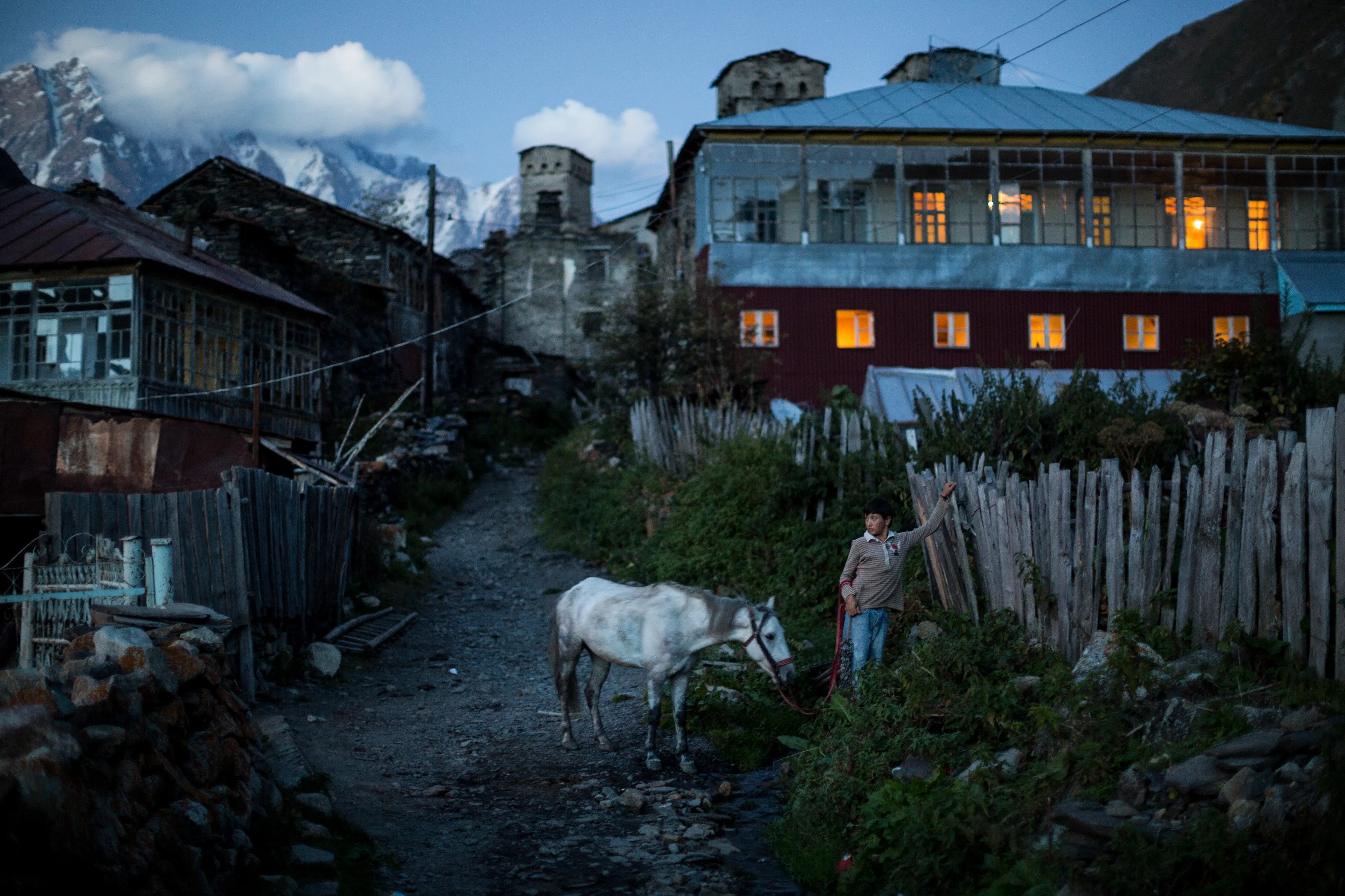 A boy puts away his horse as the blue of night closes in on the village of Ushguli, below Mt Shkhara, in Svaneti, Georgia. High in the mountains of Georgia, between the breakaway republics of Abkhazia and South Ossetia, in a valley surrounded by 5,000-meter peaks, is Svanetia (Svaneti). The old men of this valley told me that the Svans are the descendants of Sumerian slaves that escaped their bondage and fled north to the Caucasus, where they worshiped the sun god Lile’. Because of this, in all my journals, I called them: “The Children of the Sun.” I stumbled into Svaneti with a hand drawn map and a handful of slide film, and no vision for storytelling, in 1998. But over the course of 107 rolls, taken in three journeys over three years (from 1998-2000), I learned to see. The result was my first photo story, and one that would lead to my career as a National Geographic photographer. Sixteen years after that first journey, National Geographic sent me back on assignment to find my family again, and to create a feature for the magazine. This photo was taken on that return journey.  Ushguli is located at an altitude of 2,100 meters (6,900 ft) and is considered the highest continuously inhabited village in Europe. 6000  ×  4000 pixels