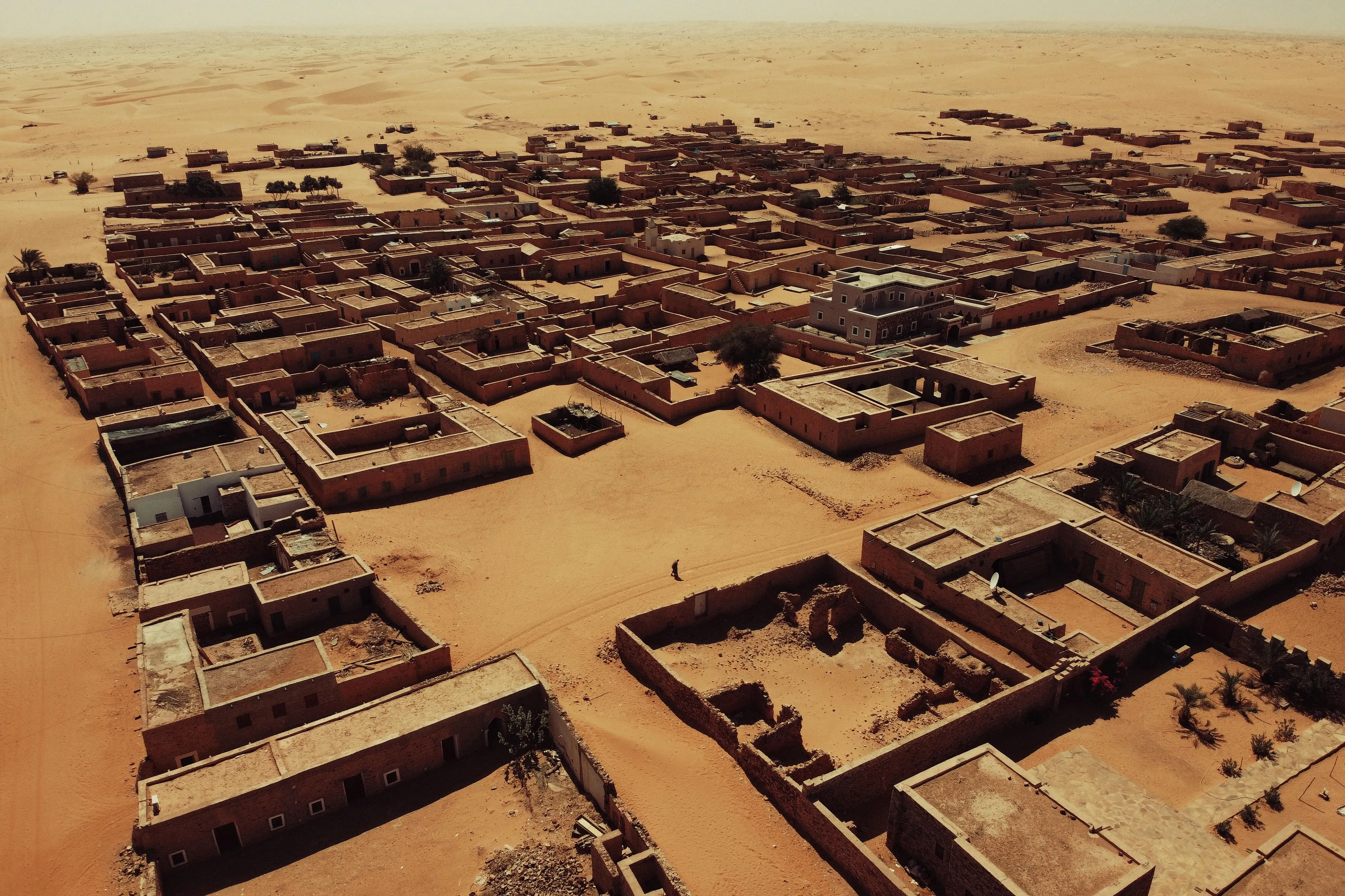 Resilience from the Latin resiliens means, in physics, the property of materials to resist shocks without breaking. 
This is where its human meaning takes its cue. Resiliens, tells the story of the relationship between man and the harshest of the environments, the desert. 
The ability of man to live, survive and adapt to the extremes of his possibilities.
In its simplicity as in its visual impact, the shot is also a reflection of my life, made up of continuous fights and challenges despite adversity and failures, the ability to be scratched and never give up.
Resilience as the basis of something greater. Not just resistance but going beyond robustness; not merely withstand a shock but actually improving because of it. 
The ability to absorb the inclements of life and make them one's own to thrive through it.
Founded in the 13th century as the center of numerous trans-Saharan trade routes, Chinguetti is a ksar in Mauritania, a medieval trading center, today severely threatened by the advance of the desert; part of the city has already been swallowed by it. A world heritage site, the city is locally considered to be the seventh holy city of Islam and is home to five of the oldest still operating libraries on the planet.
The man who walks through the streets of an ancient city that slowly sinks into the sand represents the tenacity of mankind in the face of every adversity and its resistance to surrender.
Resiliens.