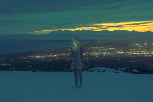 Anchorage Alaska, 2022. 

A woman stands in snow above a valley filled with city lights. The sky, rich in the colors of dusk, mirrors the pattern and hue of the lights below. The woman is blindfolded, yet seems to also be looking down at her phone, which illuminates her face softly. She holds an orb in her left hand. 

 When you look over the edge of a mountain, and notice where the sea meets the land and the mountain tops meet the sky, do you also notice where the streets end and the trees begin? Where the hand ends and the phone begins?