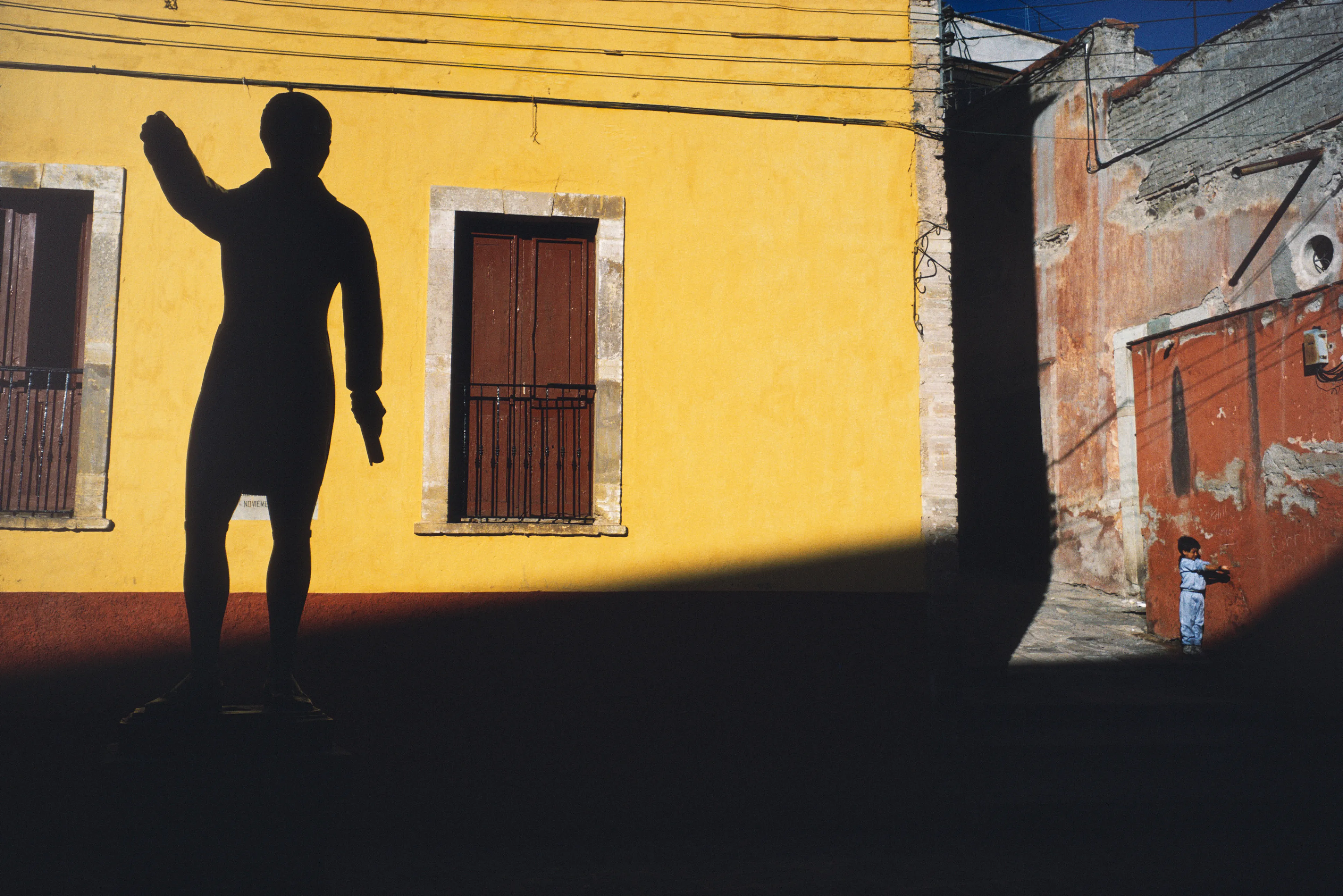 From his first visit in 1975, he was immediately captivated by the vibrant life in the streets of Mexico. While his early black and white photos often feature stark, striking graphics, his foray into color photography starting in 1978—inspired by the light and color of the Mexican streets—marks a turning point in his work.

Throughout his work from Mexico, his rich compositions create compelling visual complexity—‘La Calle’ features strong, saturated colors, which evoke haunting, enigmatic scenes of the everyday. “I work extremely intuitively. I wander, I respond,” he says. “Am I aware of certain elements rationally at times? Sure. But I think that often when I am more aware of them, it usually means that the picture falls flat.”

Webb somehow manages to capture these surprising scenes, which add an otherworldly poetry to the genre of street photography.