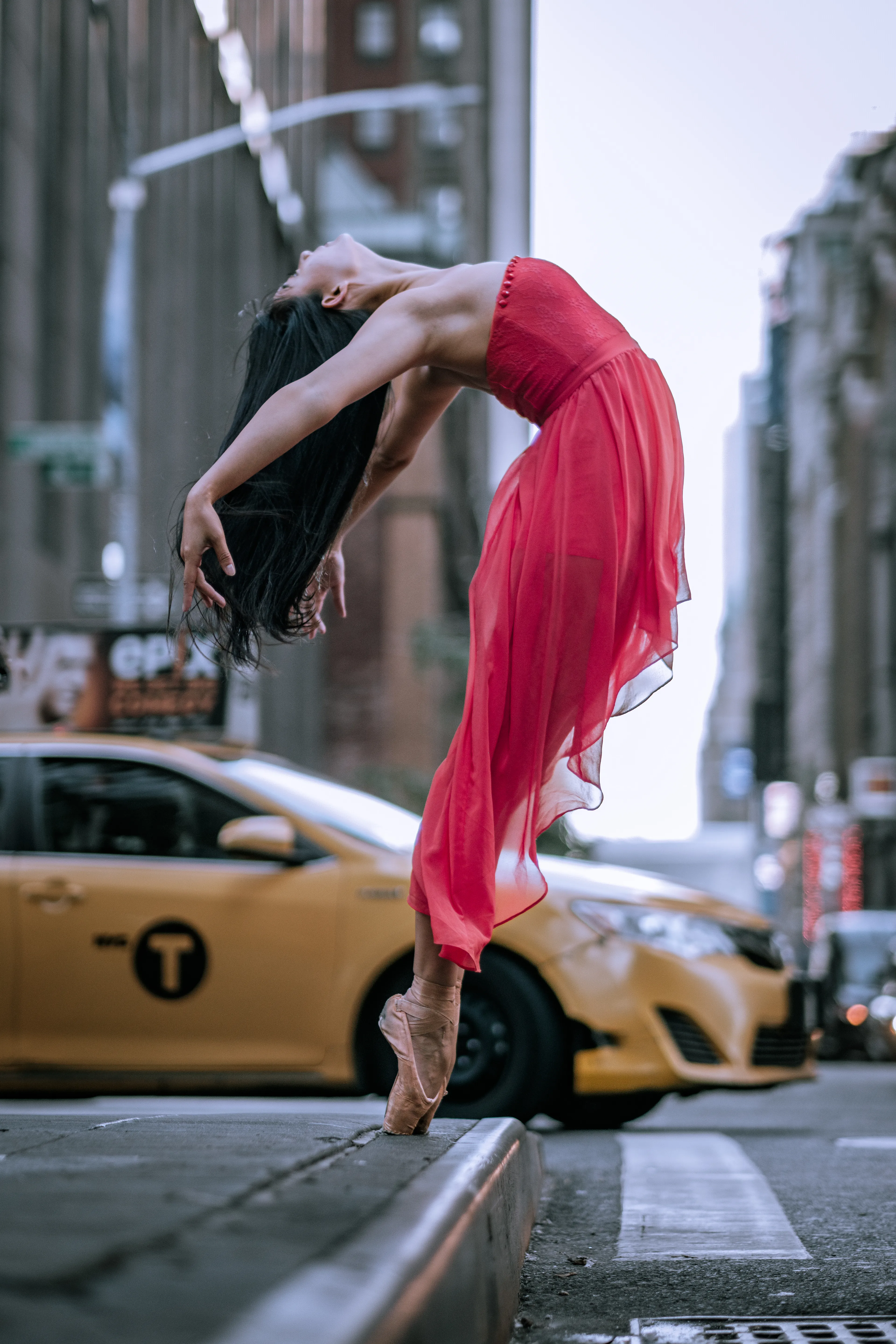 Photographed on September 15, 2015. A dancer strikes a breathtaking pose, arching backward while leaning forward on the edge of a New York City sidewalk curb. The contrast between her poised form and the bustling urban backdrop of Midtown Manhattan creates a compelling juxtaposition, portraying a decisive moment as the dancer's balance hangs in a state of suspension.