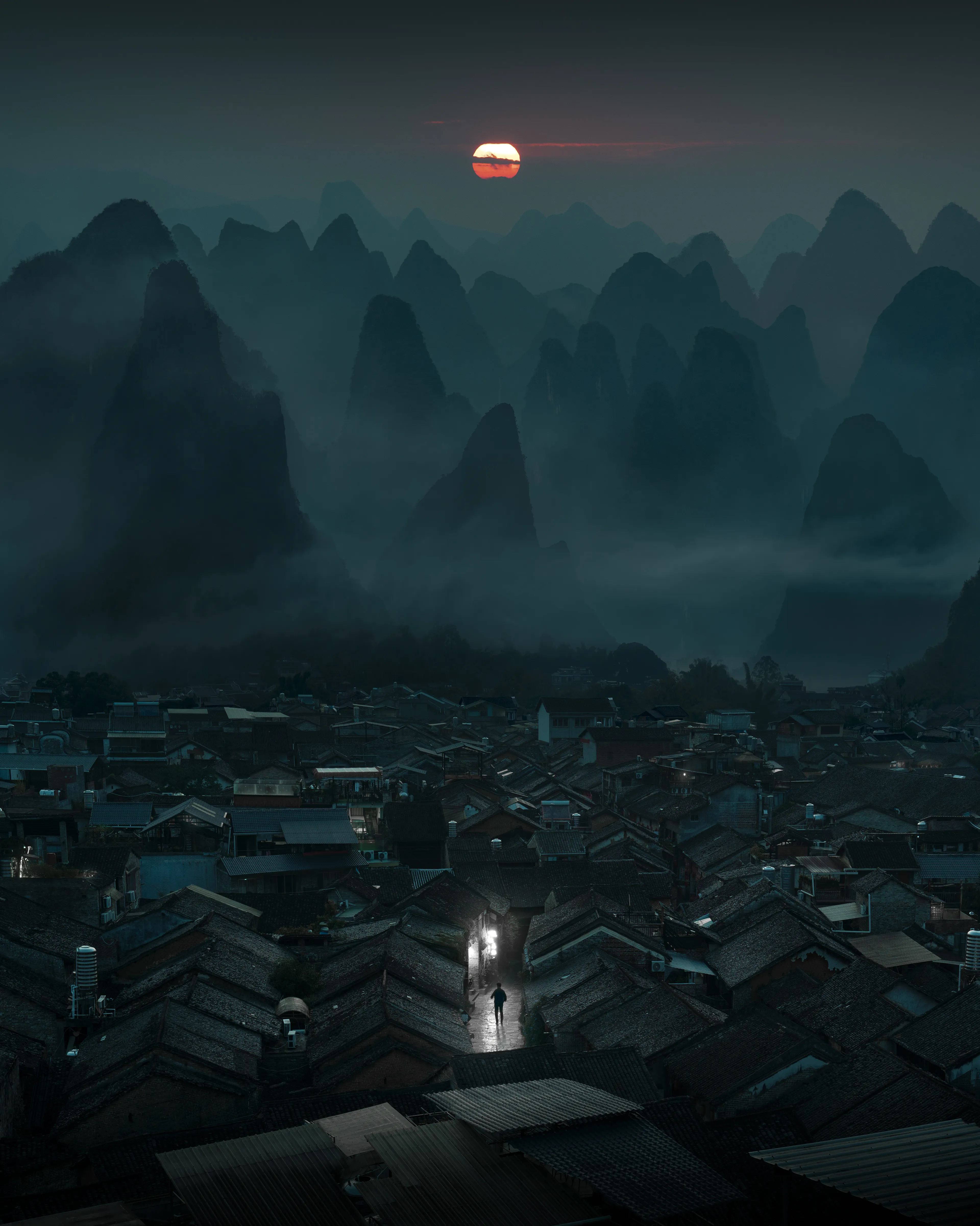 The Path of Qi is a composite created from different scenes captured around China in November 2023, blending imagination and lived experiences. The artwork immerses the viewer in the quiet energy of a secluded village, nestled within the mist-covered karst mountains.

Captured during blue hour, just before the first light of dawn, the village is still, suspended in the calm between night and day. A solitary figure appears, walking along a quiet stone path, their movement soft yet deliberate as they enter the scene. The bright lanterns that light the way symbolize Qi, the invisible force that sustains and nourishes life, casting a subtle glow in the pre-dawn stillness. The village, too, seems to hold its breath, waiting for the first signs of life.  
 
The jagged mountains, looming over the village, reflect the ruggedness of the human experience; sharp, fractured, yet ever-present, mirroring the complex and interconnected flow of Qi.

The village, with its tightly clustered houses, appears almost as if it were a miniature landscape, mirroring the mountains in their compactness and closeness. Just as the mist encircles the landscape, so too does Qi intertwine with every moment of existence, felt in the air, the land, and the people. 

The figure’s solitary journey becomes a reflection of introspection and purposeful solitude. It is not an act of isolation but one of connection, a quiet search for alignment with the natural world and its energies. The figure’s presence serves as a reminder of the deeply personal yet universal journey of finding balance within oneself amidst the vastness of existence.

The Path of Qi’s medium and story mirror each other. The careful assembly of disparate elements reflects the Taoist principle of balance of how all things, even those that appear separate, are ultimately part of a greater, seamless whole. Just as Qi flows through every aspect of life, the creative process behind this piece mirrors that constant flow of energy, where every photograph plays its part in creating a unified, cohesive journey.